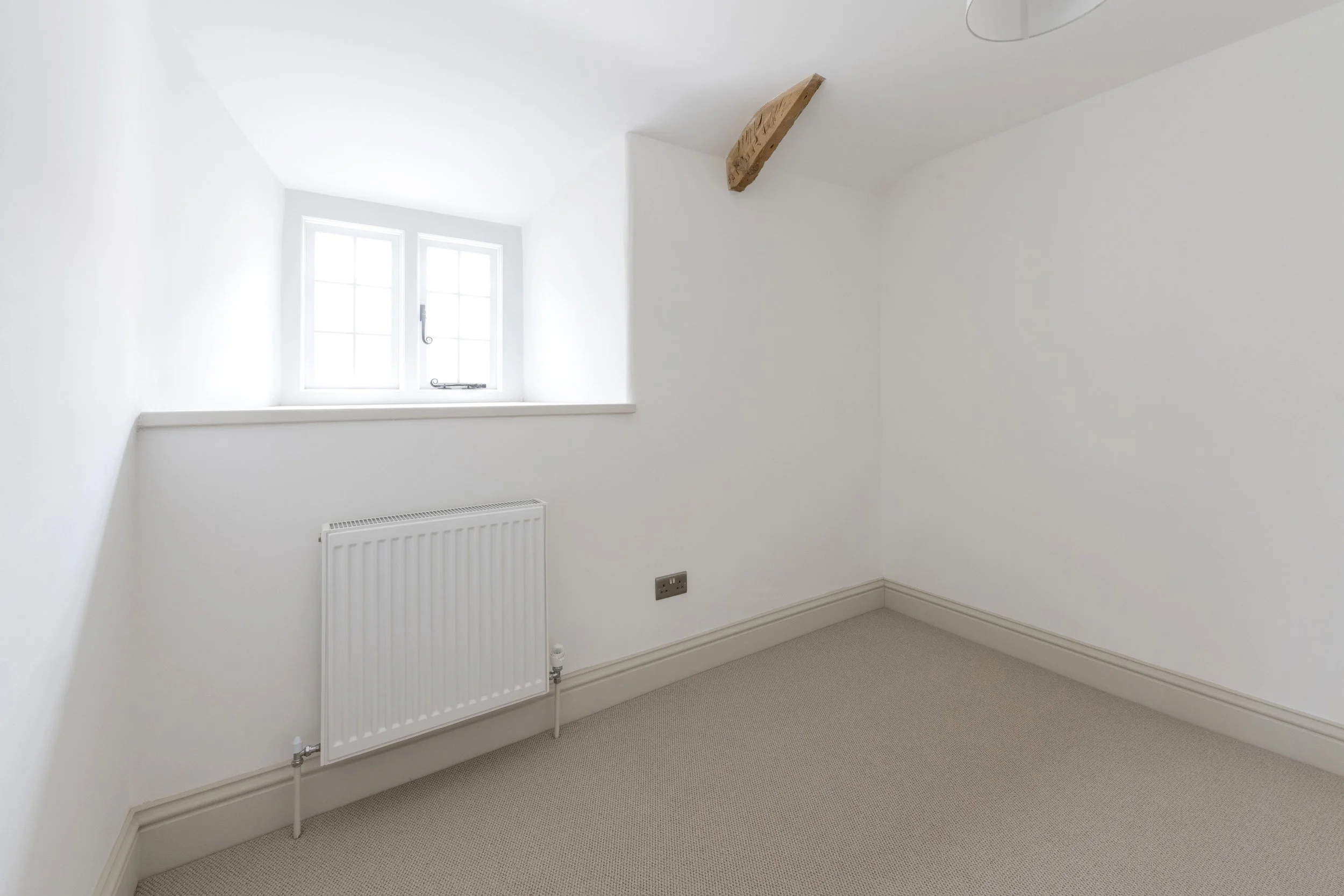 Empty white room with a small window, beige carpet, and a radiator under the window, featuring a wooden beam on the ceiling.