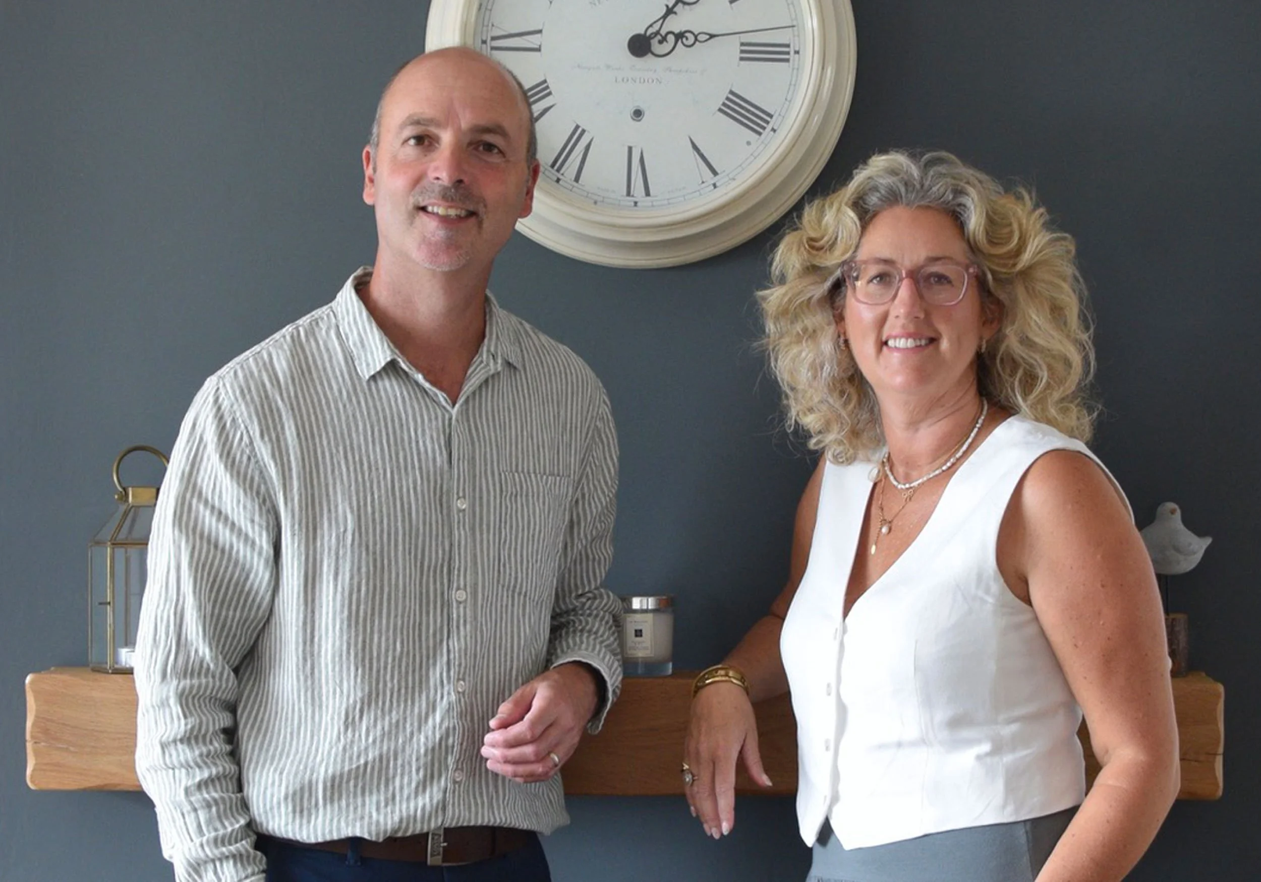 The owners and founders of The Home Staging Specialists, standing side by side indoors in front of a dark wall with a large clock. The man is wearing a striped button-down shirt and the woman is wearing a sleeveless white top, glasses, and jewelry.