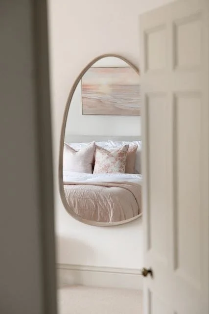 A bedroom seen through a partially open door with a large oval mirror reflecting a bed and decorative pillows.