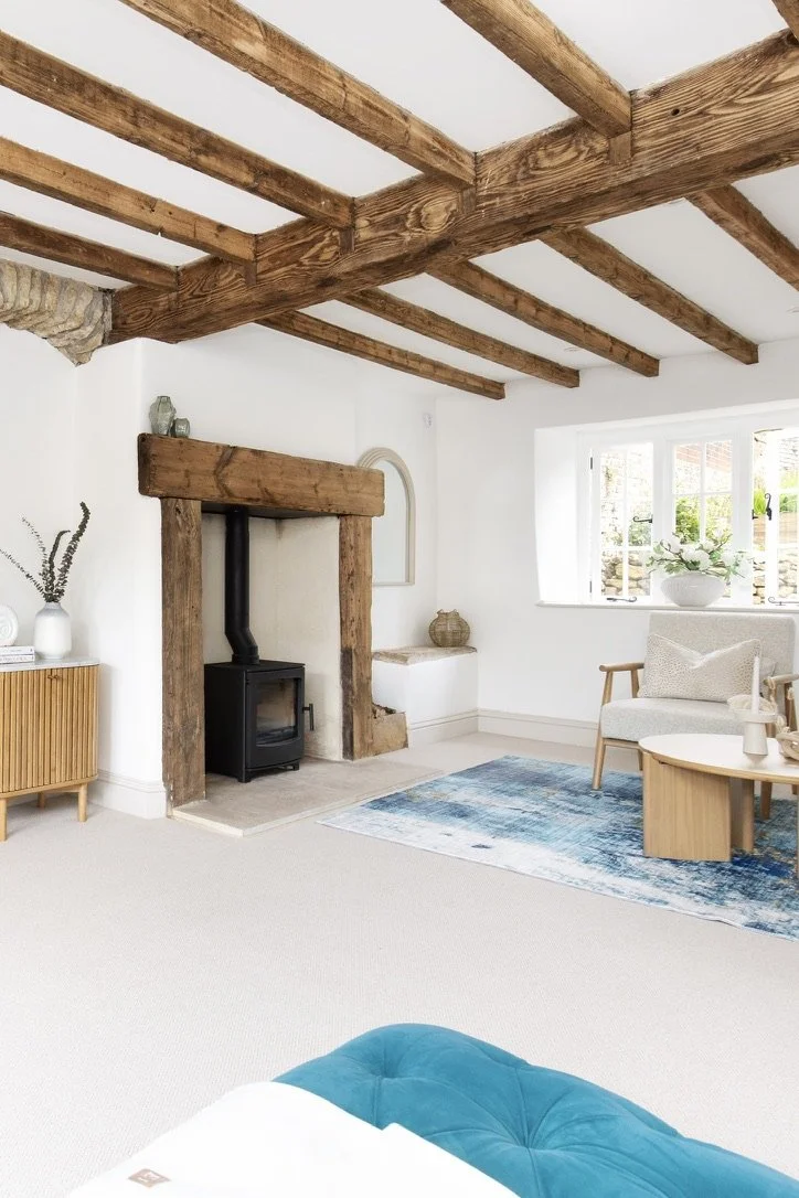 Living room with wooden beams on the ceiling, a fireplace with a wood mantel, a window with white framing, a blue area rug, a cream-colored armchair, an oval wooden coffee table, a side dresser with decorative vases, and minimal décor.