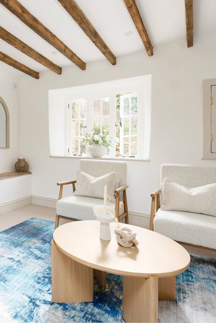 Bright living room with white walls, wooden ceiling beams, a window with outdoor view, white upholstered chairs, a light wood coffee table, and a blue patterned rug.
