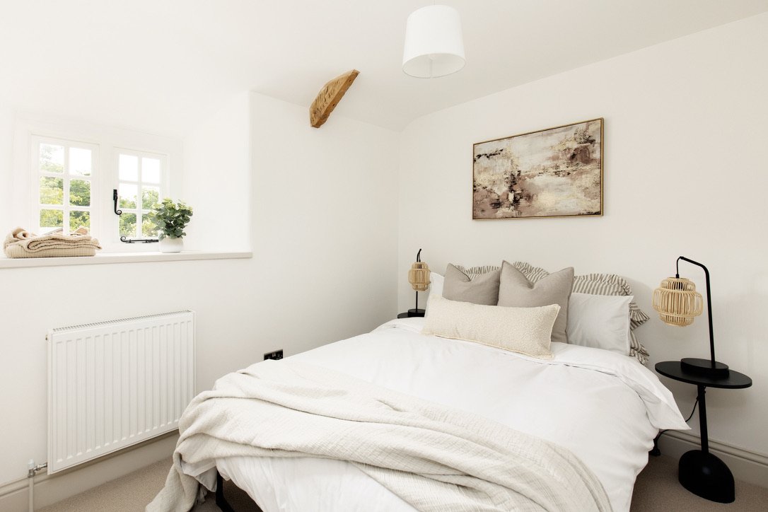 A minimalist bedroom with a white bed, beige pillows, wall art, black bedside tables with wicker lamps, a window with a potted plant and folded towels, and a wooden beam on the ceiling.