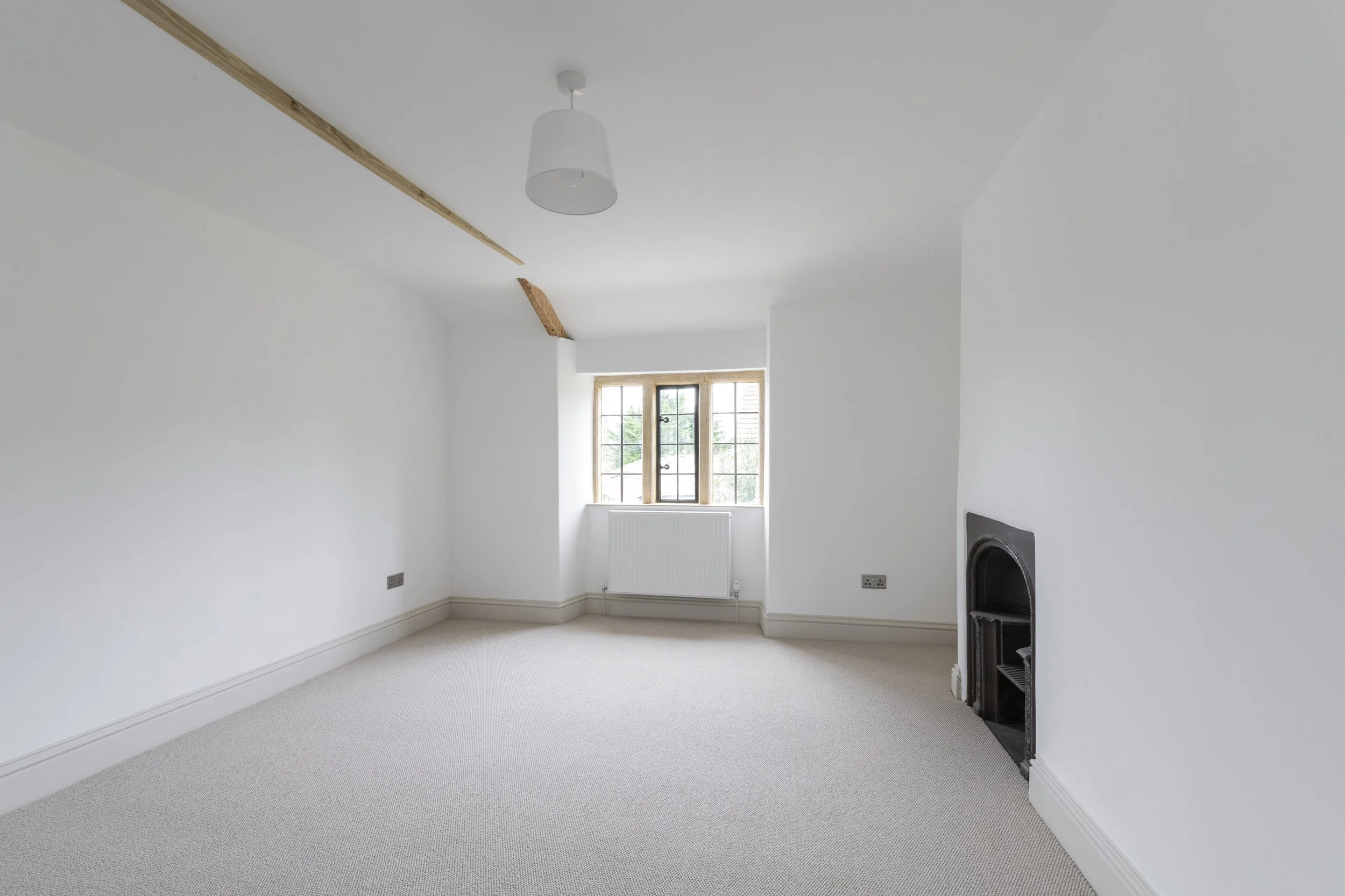 Empty white room with carpeted floor, a window, a small black fireplace, and wooden beams on the ceiling.