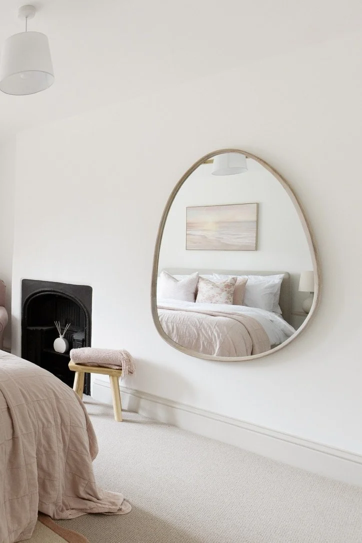 A bedroom with neutral-colored bedding, a large round mirror on the wall reflecting the bed and a painting, a small stool with a pink towel, and a black accent wall with a white vase and reed diffusers.
