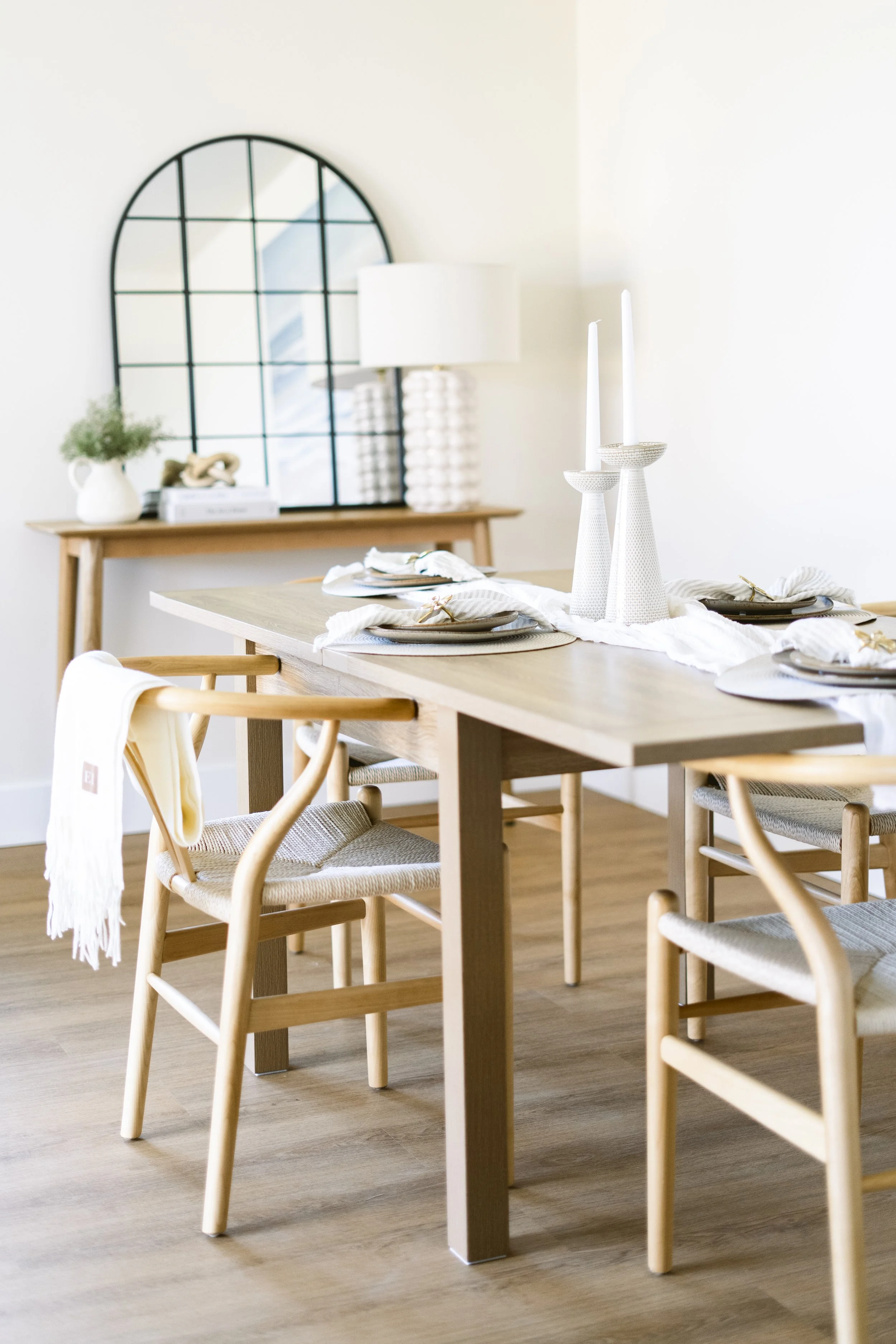 Professionally staged dining room with a wooden dining table, natural wood chairs, neutral tableware and a console table with a large arched mirror.
