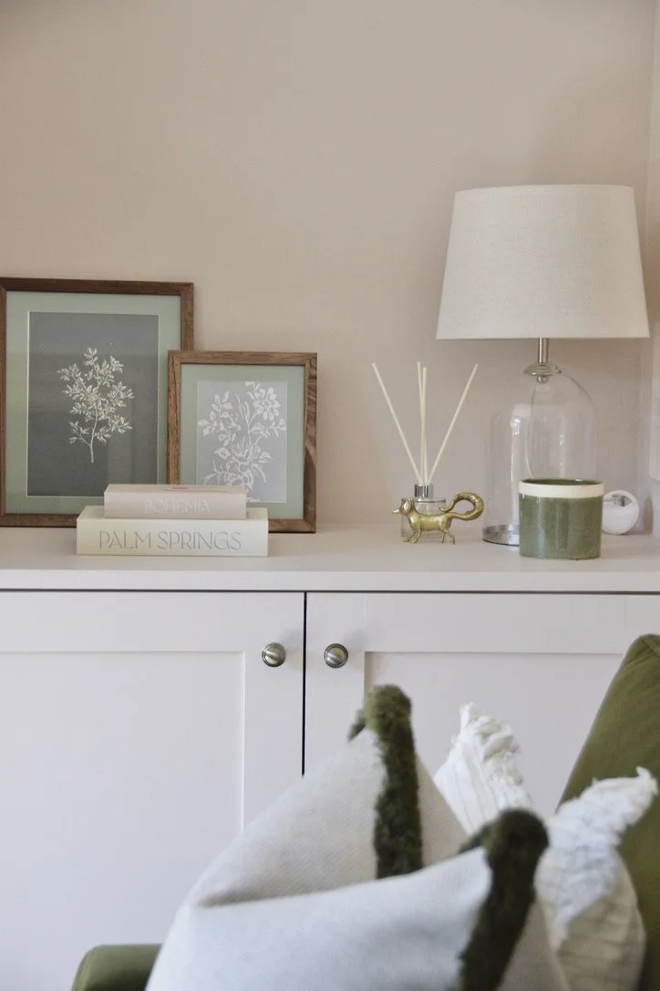 Decorative living room corner featuring framed botanical prints, books titled 'Potennia' and 'Palm Springs', a glass lamp with a white shade, aroma reed diffuser, green ceramic mug, white ceramic vessel, and part of a green chair or sofa with textile and blanket draped over it.