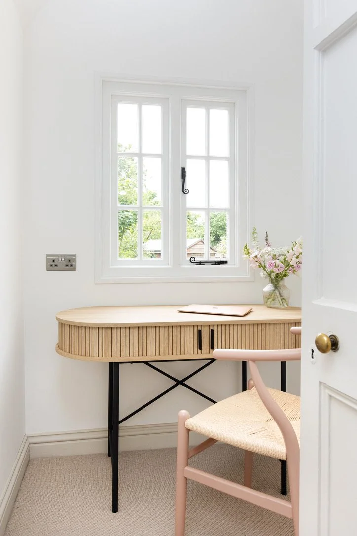 A small, modern home office space with a wicker desk, pink chair, and a vase of pink and white flowers on the desk, located near a window with white framing, in a bright room with white walls and beige carpet.
