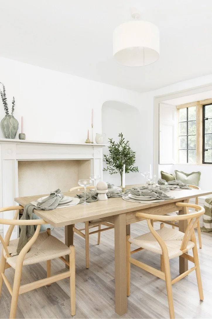 A dining room with a wooden dining table set with plates, napkins, and glasses. Four light wooden chairs with woven seats are around the table. A small green potted plant is in the center. In the background, a white fireplace and a window with black-framed panes are visible, allowing sunlight into the room. The room has a minimalist, bright, and airy decor.