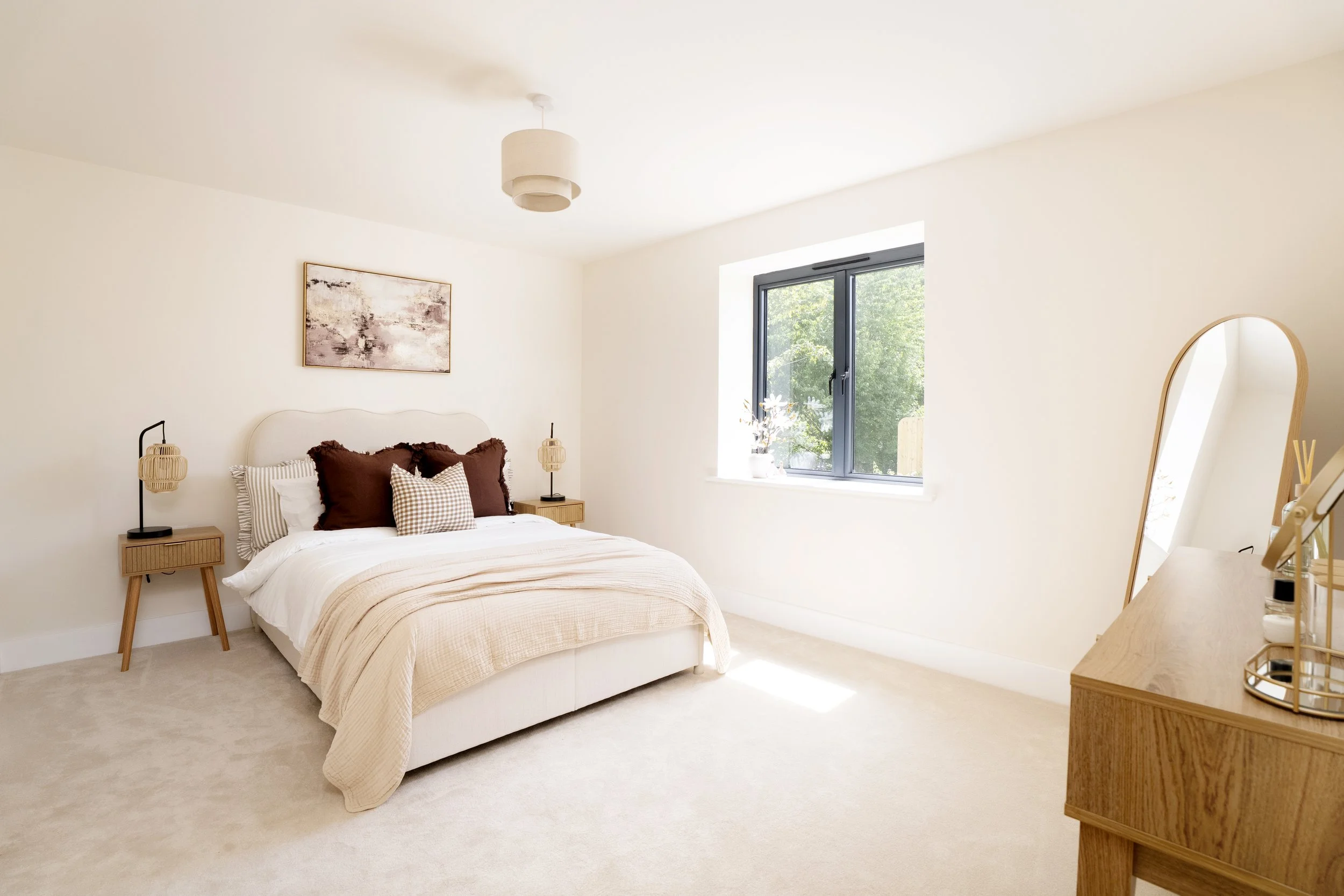Professionally staged spacious bedroom in a Somerset property with neutral upholstered bed, warm brown accents and oak furniture. Bed has a neutral headboard with brown cushions & a gingham accent cushion. Floor standing mirror in corner of room.