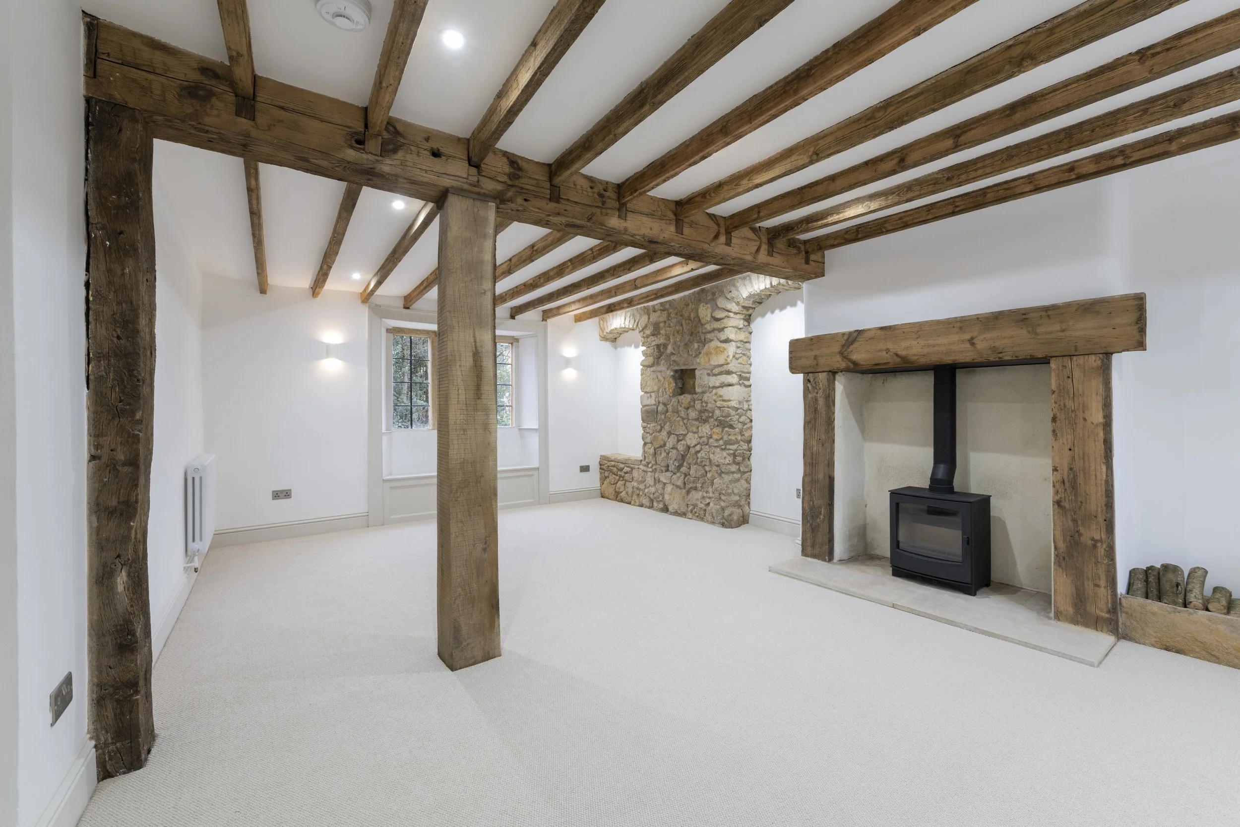 Empty living room with white walls, wooden ceiling beams, a stone fireplace, and large windows.