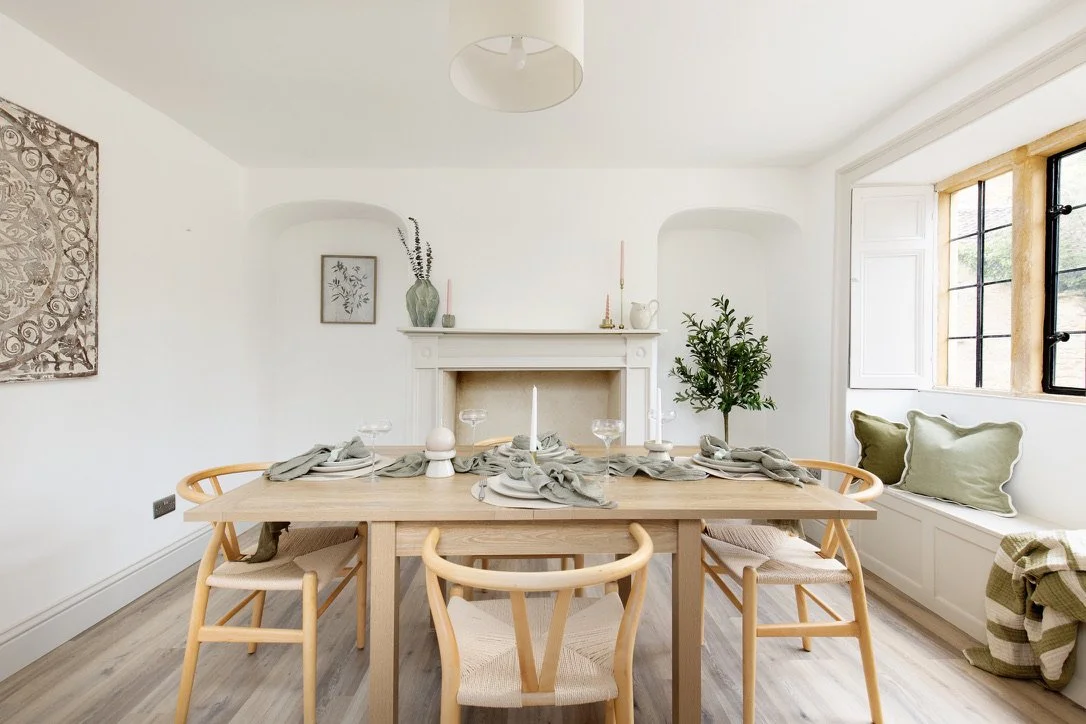 Dining room with wooden table set for six, surrounded by light wood chairs, with a white fireplace and window seat with green cushions, decorated in neutral tones.