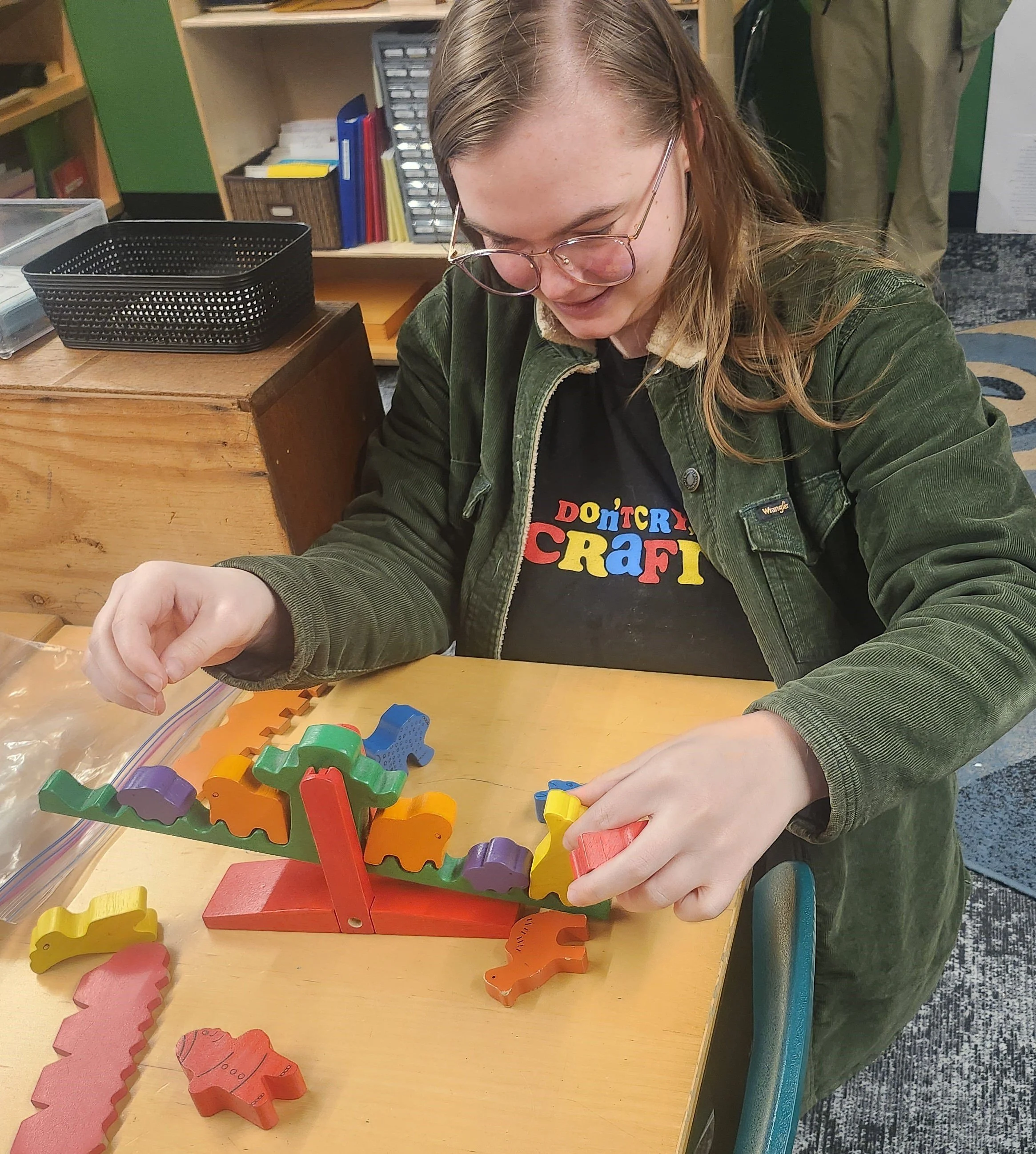 A person wearing glasses and a black shirt with colorful text is playing with colorful wooden animal-shaped pieces and a balancing game on a yellow table.