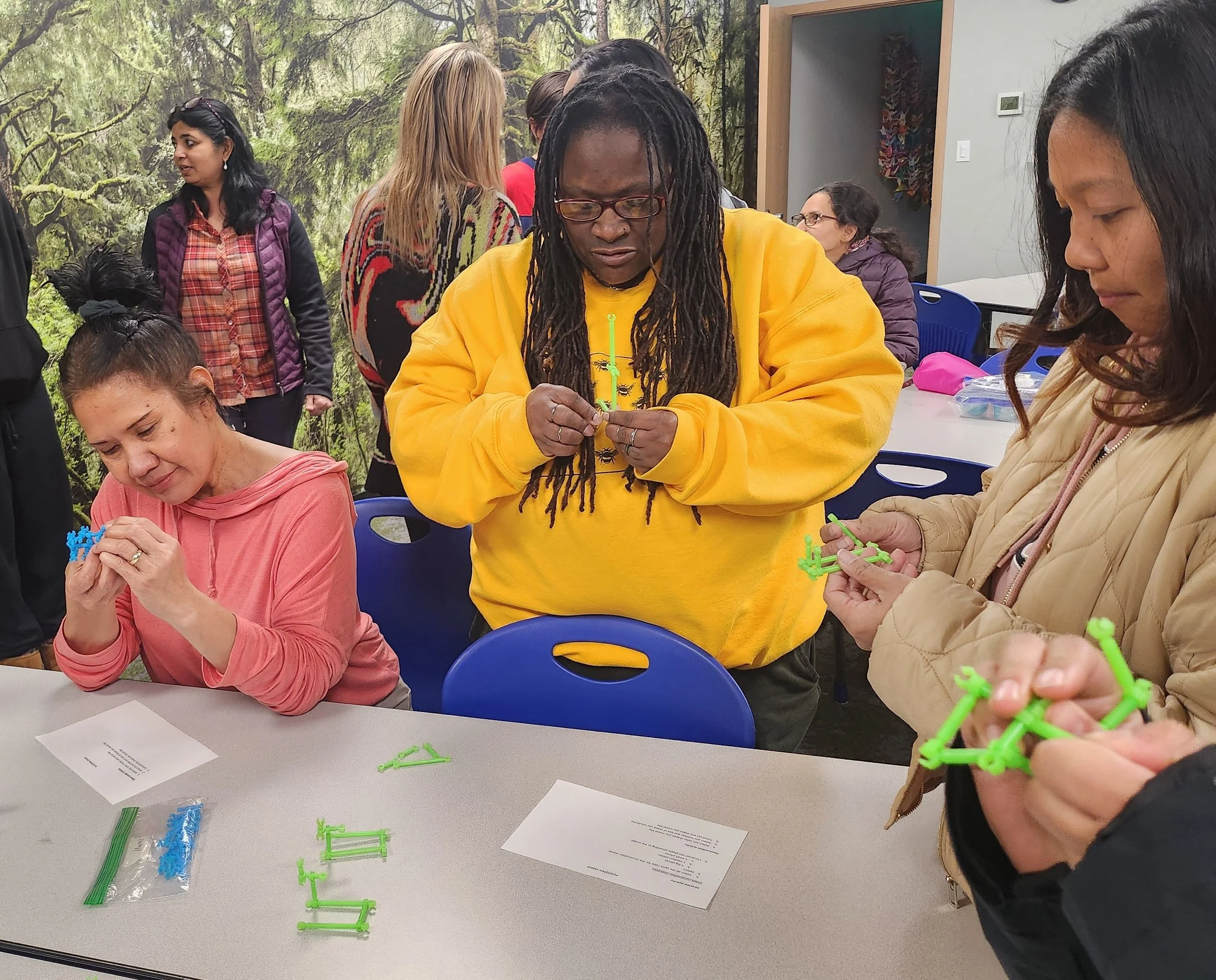 Group of people assembling small green and blue plastic models at a table in a room with forest-themed wall decor.