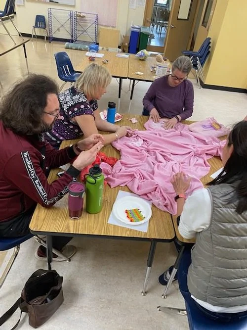 Four women sitting around a table, working on a pink blanket or fabric, in a room with chairs and tables.