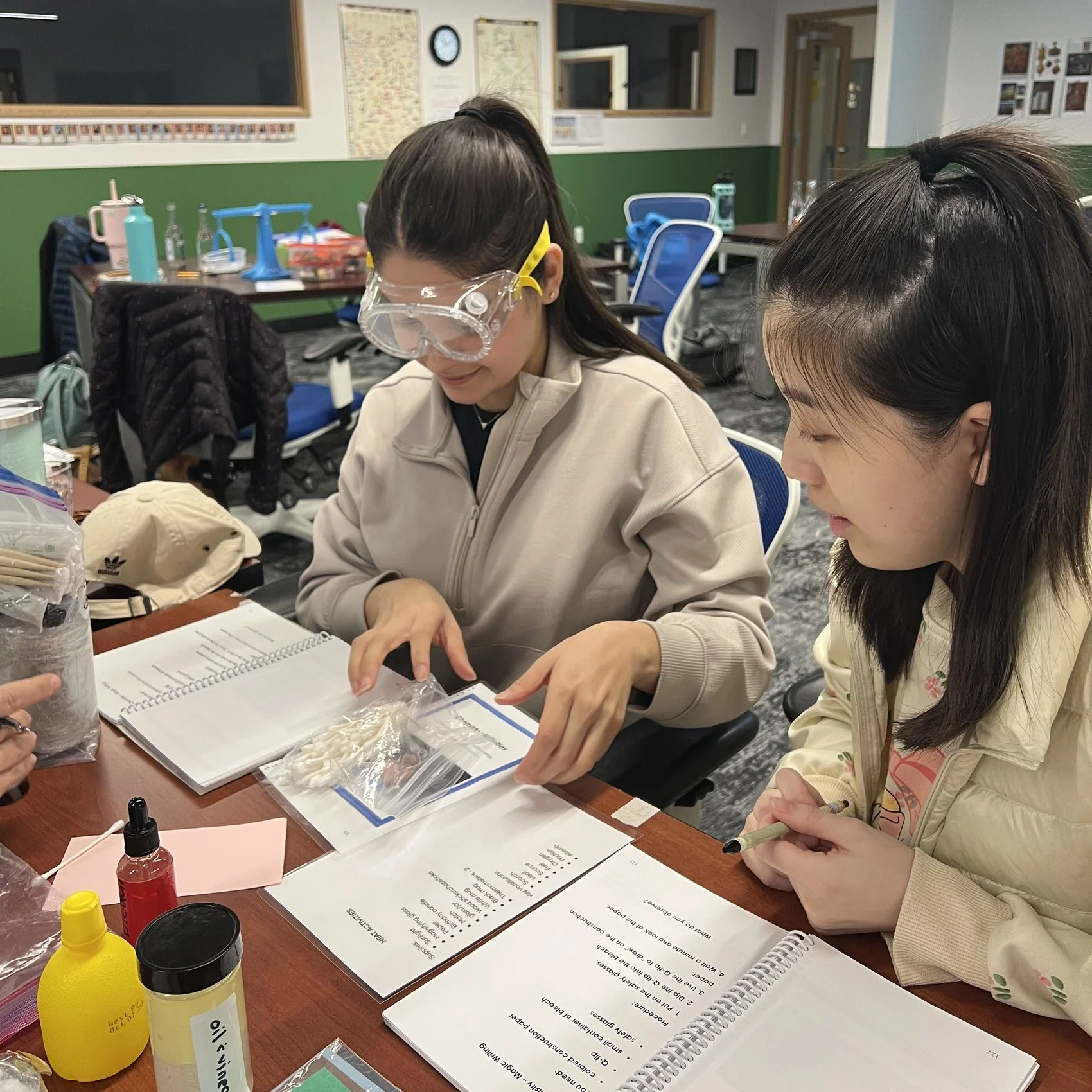 Two women sitting at a table in a classroom, engaged in a science experiment or activity. One woman is wearing goggles and a beige jacket, and the other woman has long dark hair and a light-colored jacket. The table has notebooks, a sealed plastic bag, and various small containers and supplies.