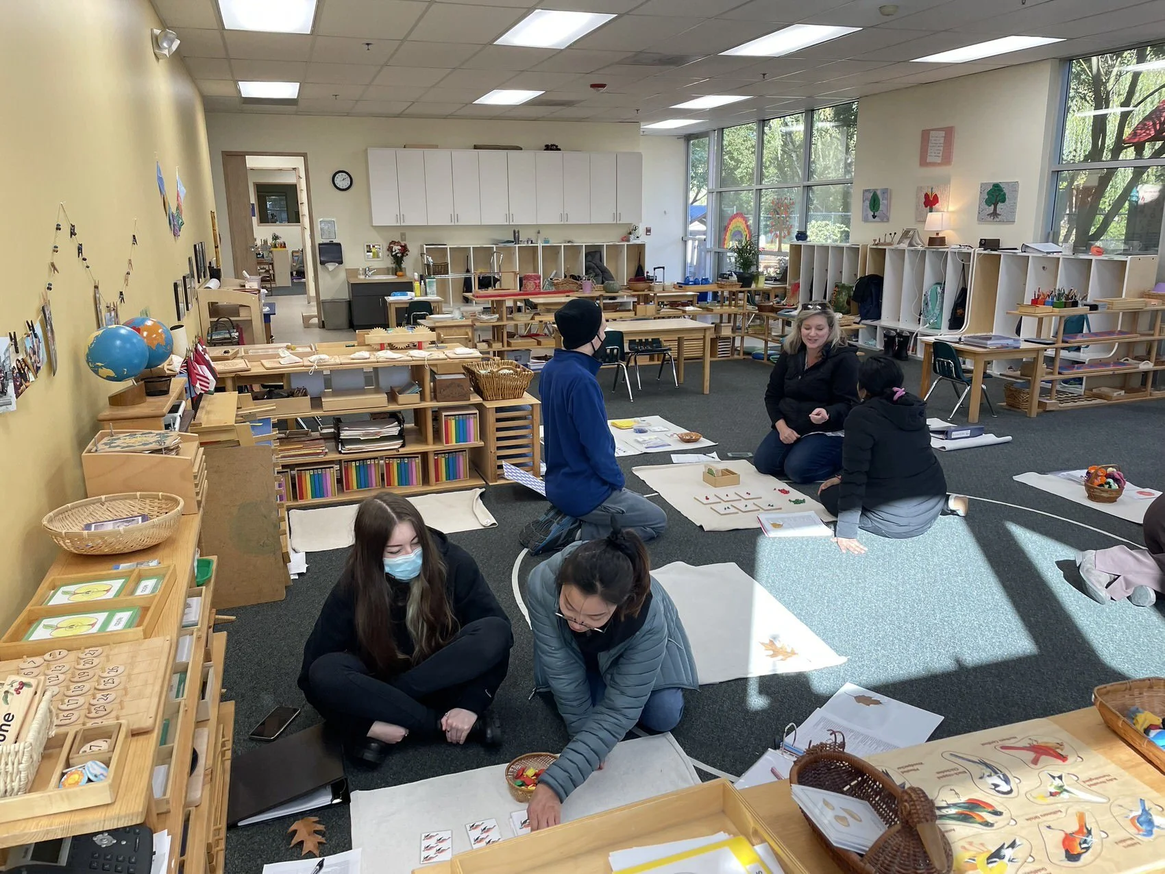 Classroom with children and teacher engaged in learning activity on the floor, with educational materials, books, and artwork displayed around the room.