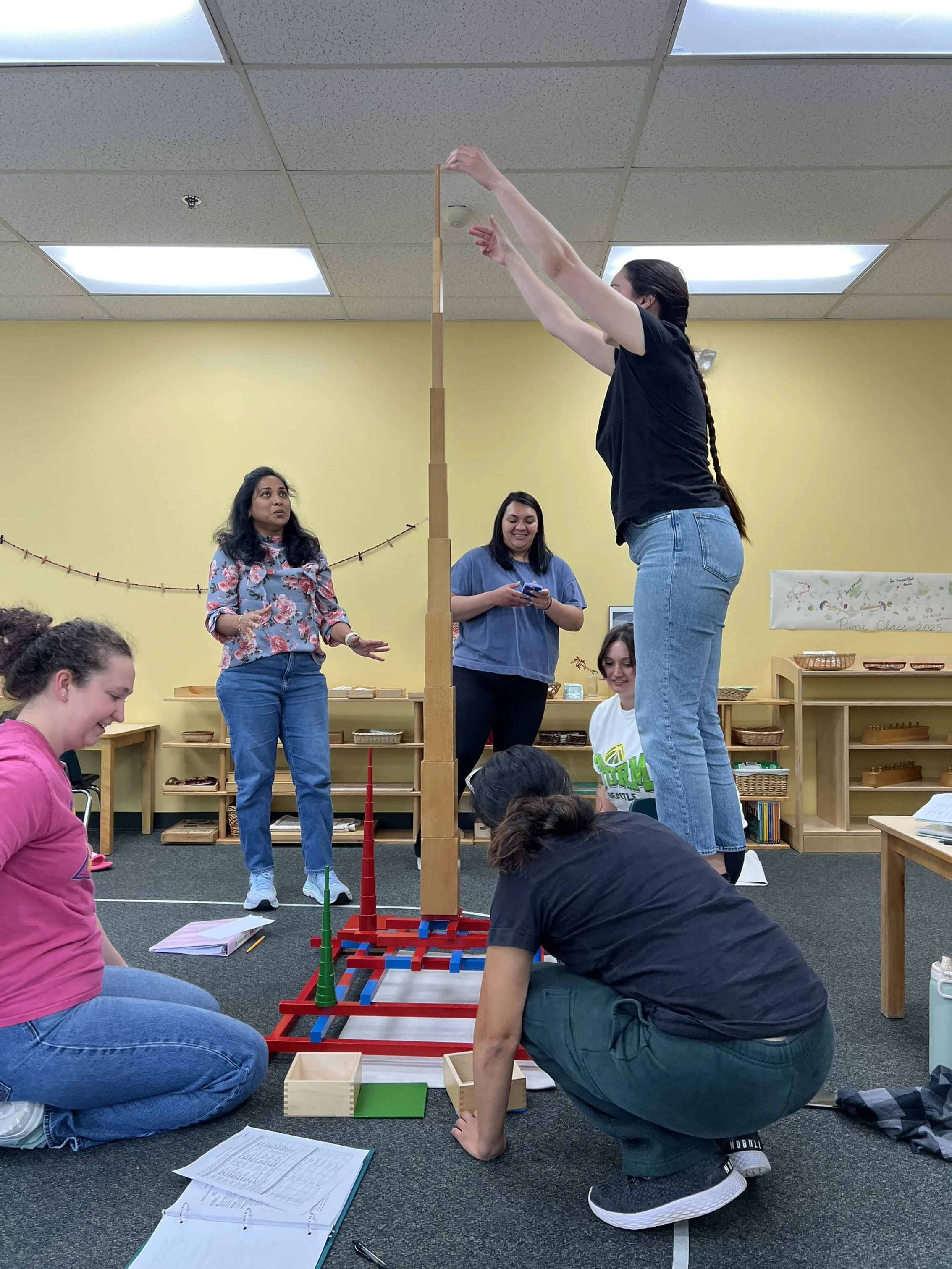 Group of women building a tall tower with wooden blocks in a classroom, with some women sitting on the floor and others standing.