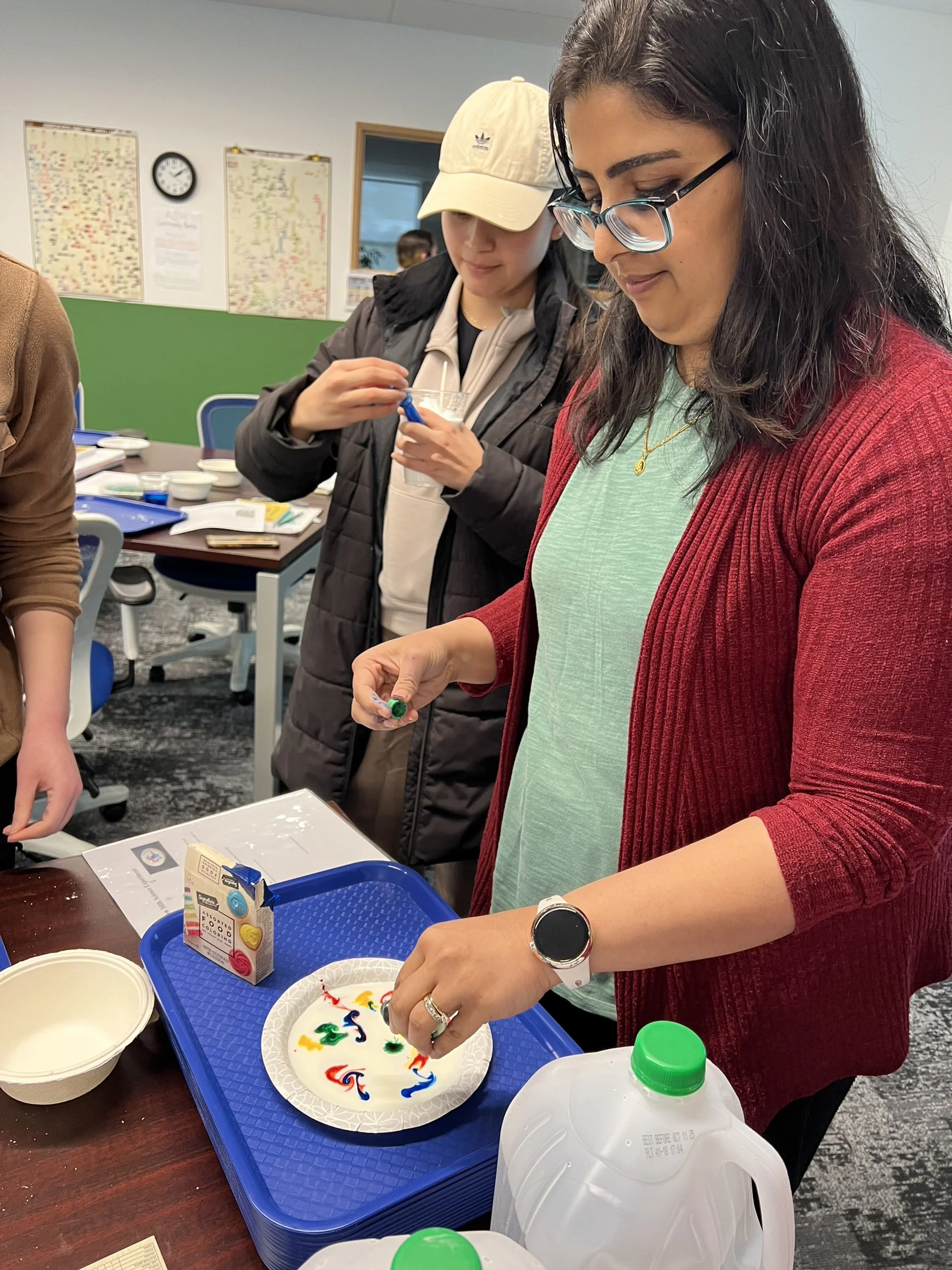 Two women participate in a hands-on activity at a table, involving colorful swirling liquids on a paper plate. One wears glasses and a red cardigan, and the other, a white cap and a dark jacket, is observing. The background shows a classroom or offic