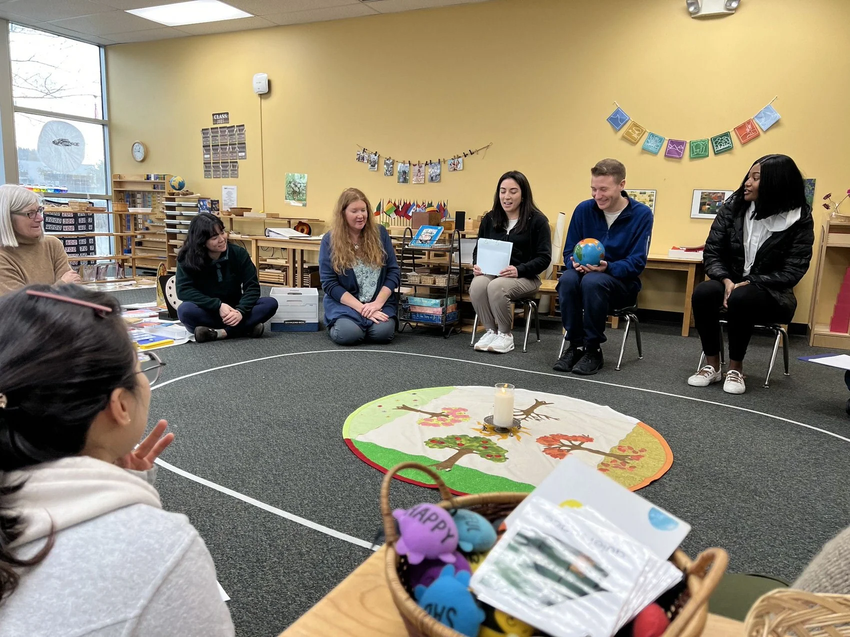 Group of people sitting in a circle on the floor and chairs in a classroom, participating in a discussion or activity. There is a decorative mat with trees and a candle in the center, and the classroom walls are decorated with colorful banners and pictures.
