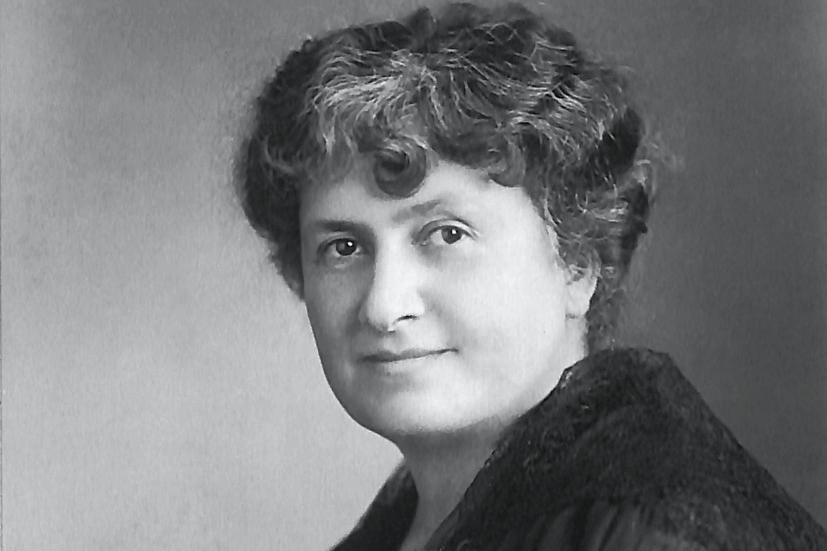 Black and white portrait of Maria Montessori with short curly hair, wearing a dark top, looking at camera with a slight smile.