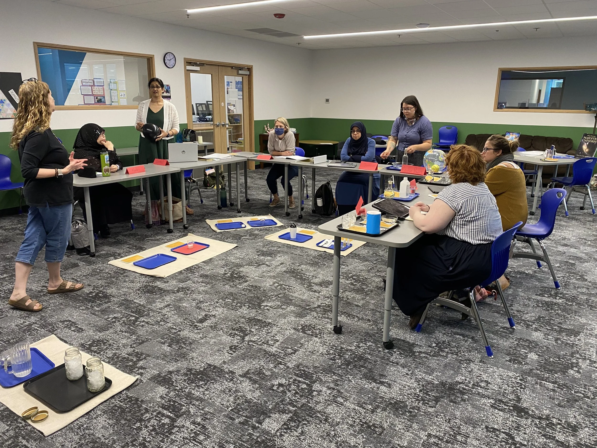 A group of women participating in an indoor workshop or class, sitting at tables arranged in a U-shape, with some taking notes. In the middle of the room, there are some display trays with jars and other items, and a woman standing and presenting. Th