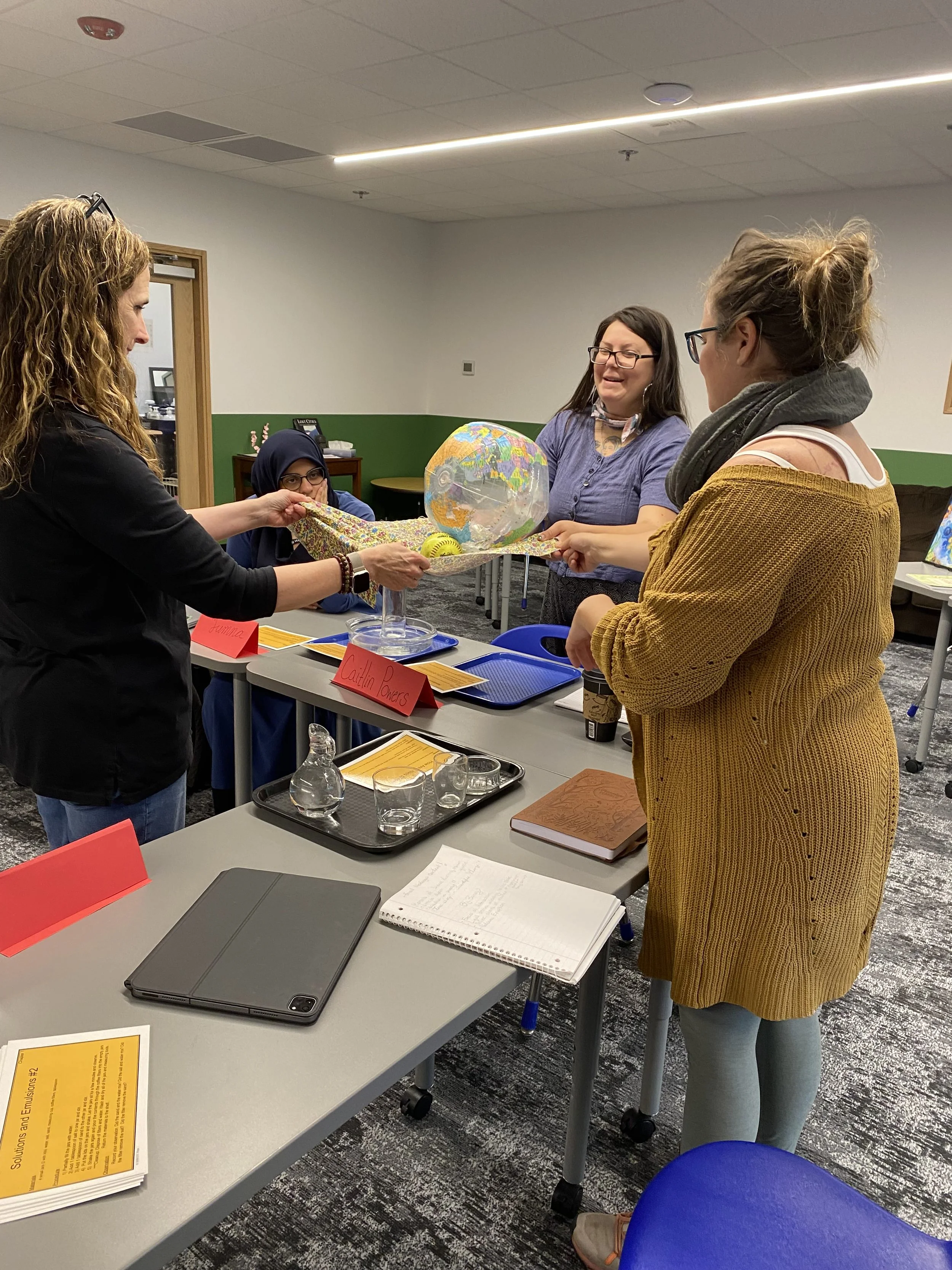 Four women are gathered around a table with a display of a globe, a yellow ball, and a colorful fabric. One woman is holding the fabric with her hand, while another woman is smiling and engaging in conversation. The table has trays, notebooks, a tabl