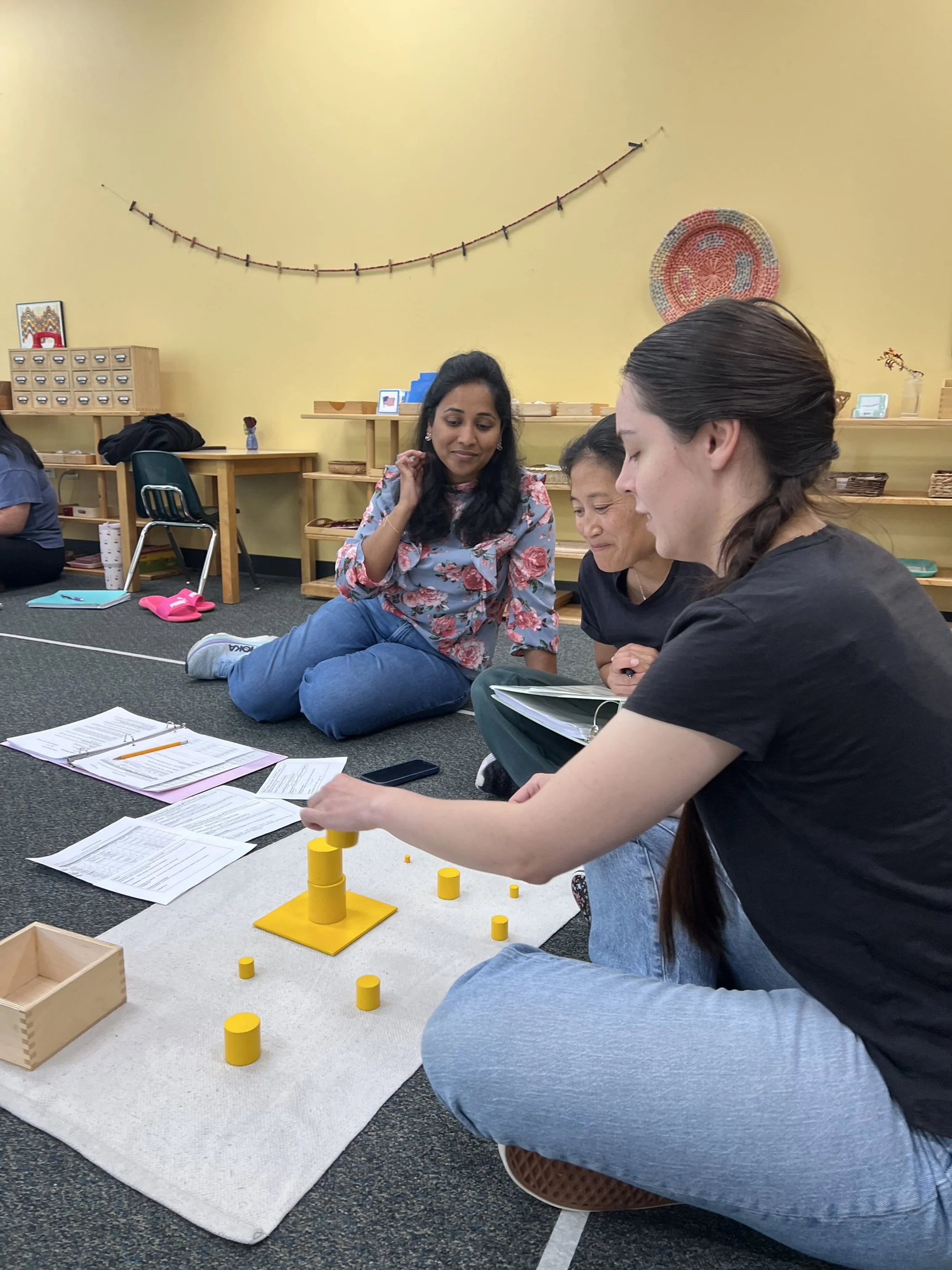 Three women sitting on the floor engaged in a conversation or activity, with papers, notebooks, and yellow cylindrical object in front of them, in a room with shelves and yellow walls.