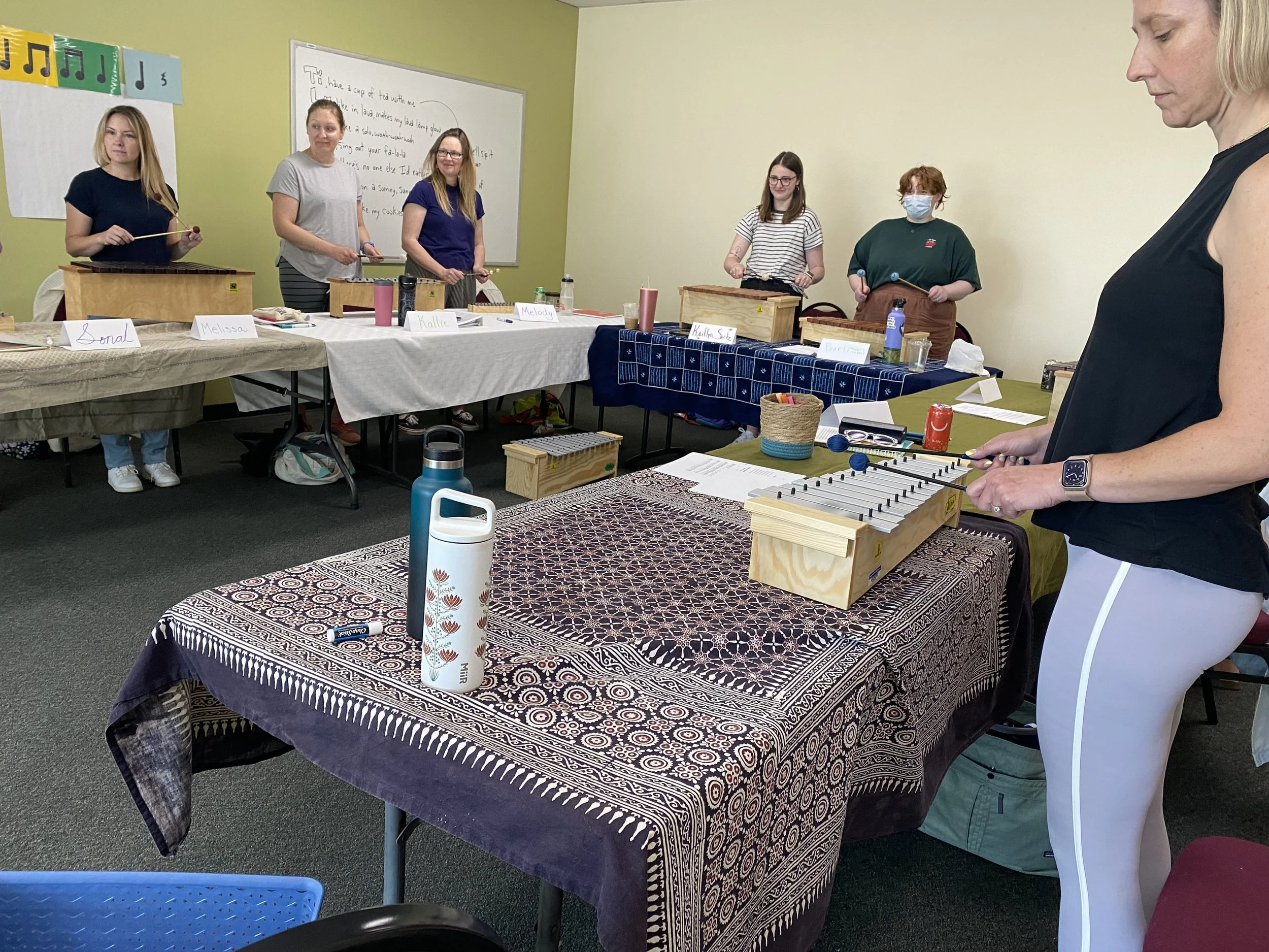 Women playing xylophones and percussion instruments in a classroom or workshop setting.