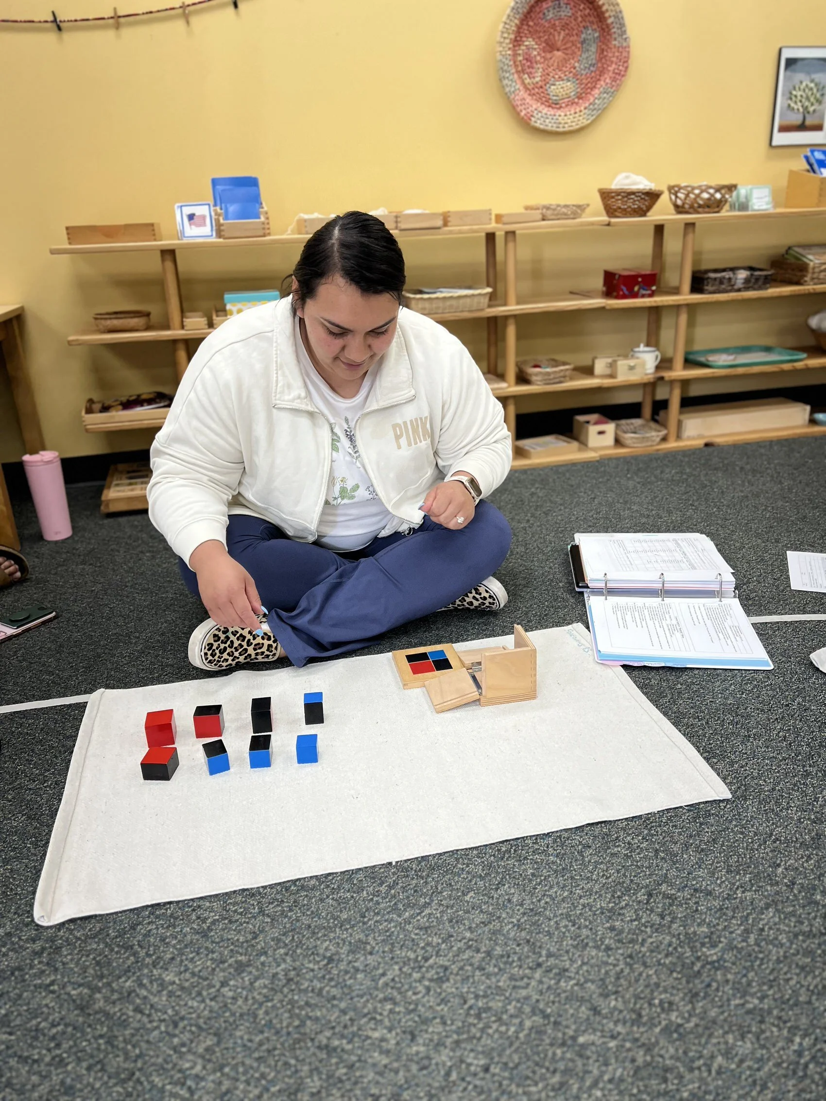 A woman sitting cross-legged on the floor with pattern blocks and an activity mat, engaging in a learning activity in a classroom setting with shelves, baskets, and decorative items in the background.