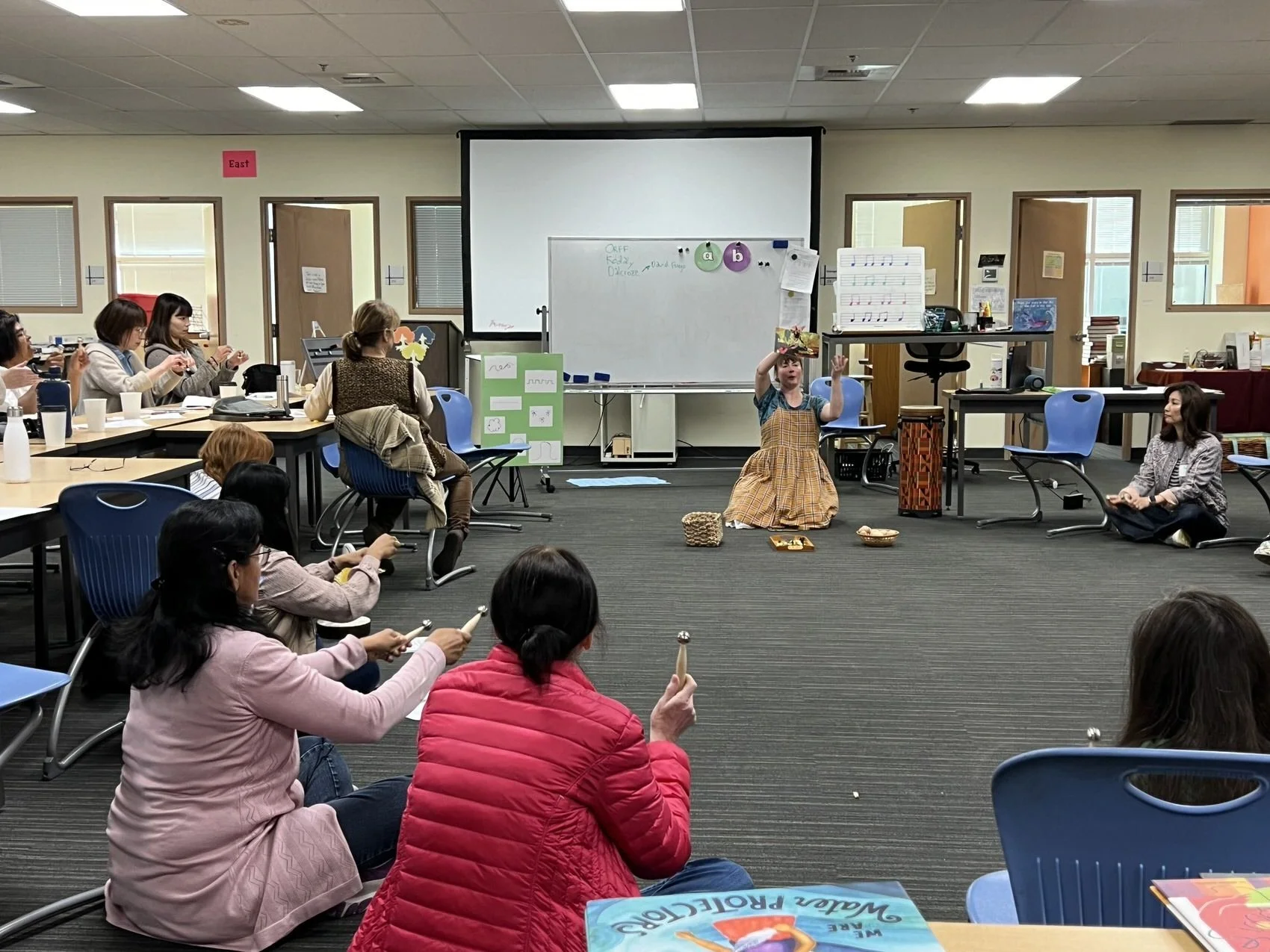A woman in a plaid dress sits on the floor in front of a whiteboard, holding a book above her head while children and adults watch in a classroom or meeting room. The room has chairs, tables, and educational materials.