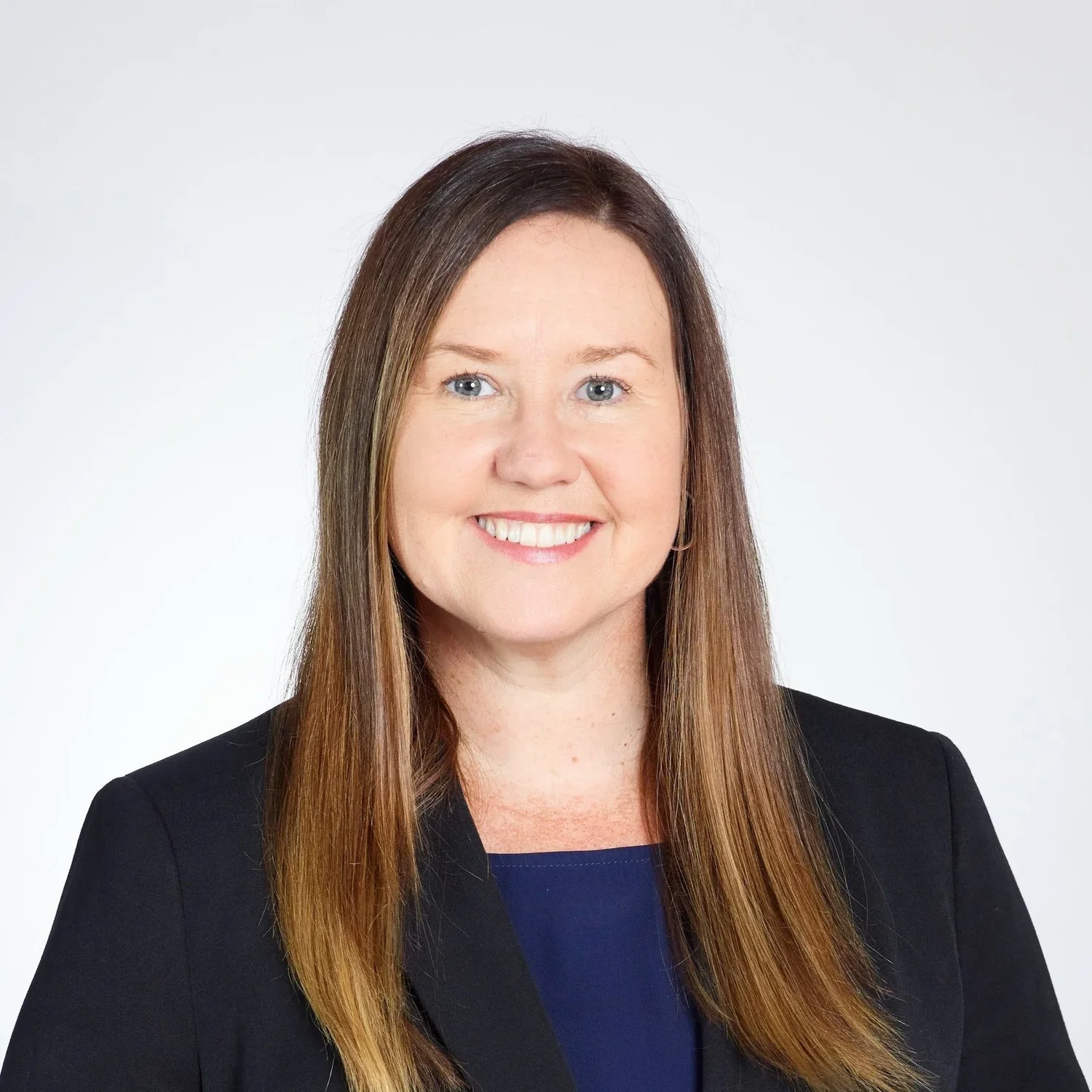 Professional woman with long brown hair, blue eyes, smiling, wearing a black blazer and navy blue top, against a plain white background.