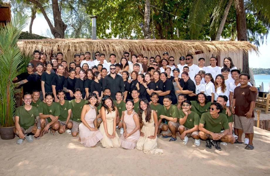 Large group of people gathered outdoors at a beachside location, with a thatched-roof hut and trees in the background, posing for a photo during daytime.