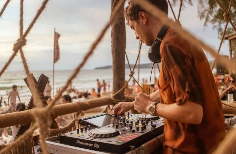 A DJ wearing a brown shirt and watch is playing music on a Pioneer DJ controller at a beach party with people in the background, some on the beach and others in the water during sunset.