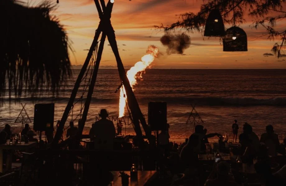 People gathered on a beach at sunset with a bonfire and string lights, watching a fire-breathing performer over the water.