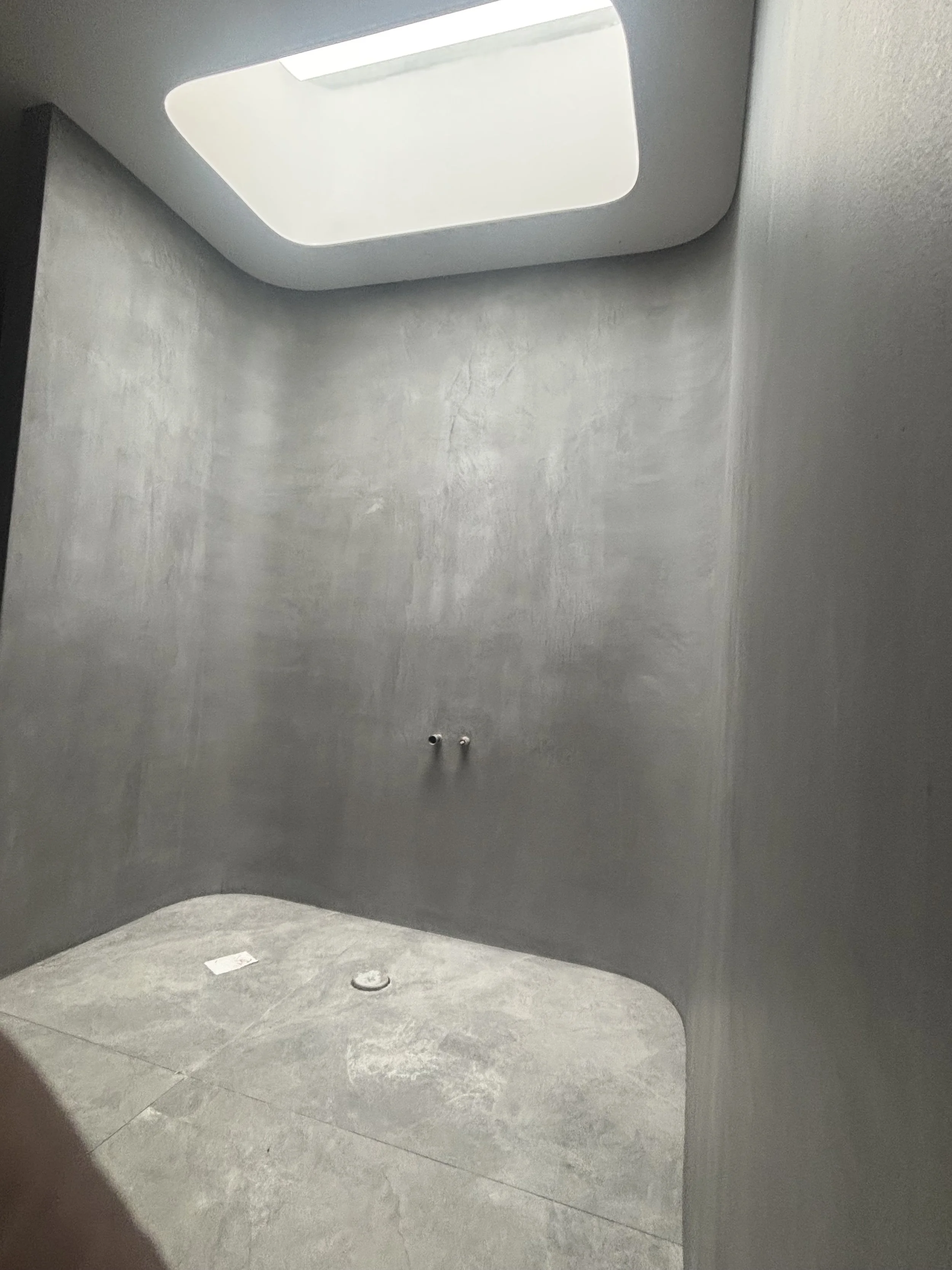 Interior of a modern, unfinished shower with gray walls and floor, a skylight, and plumbing fixtures.