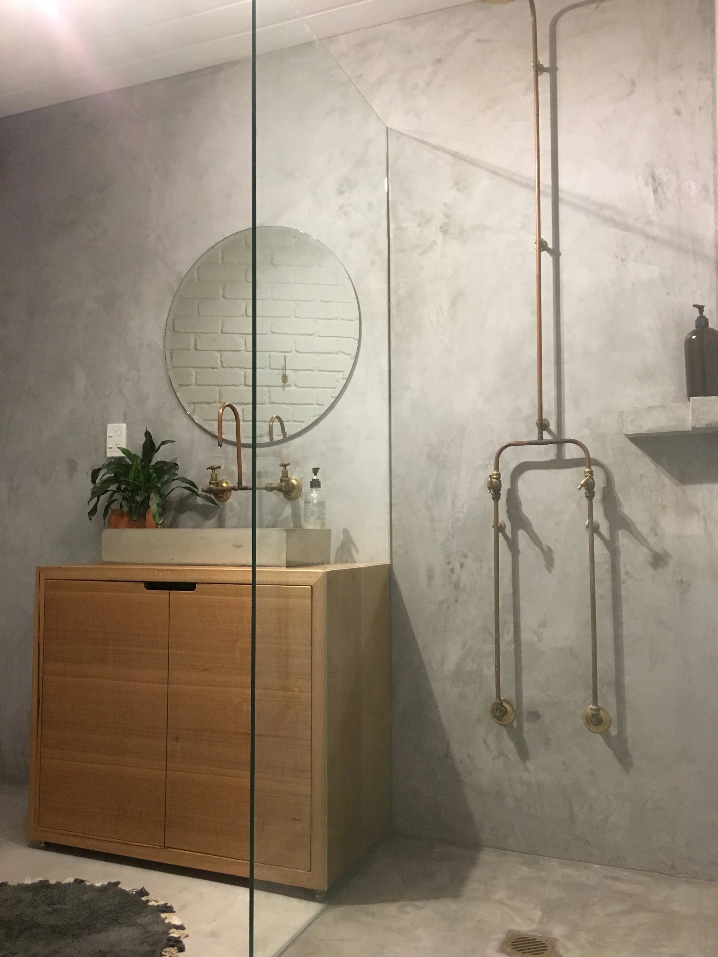 Modern bathroom with concrete walls, a wooden cabinet with a concrete sink, a round mirror, a potted plant, and exposed plumbing fixtures on the wall.