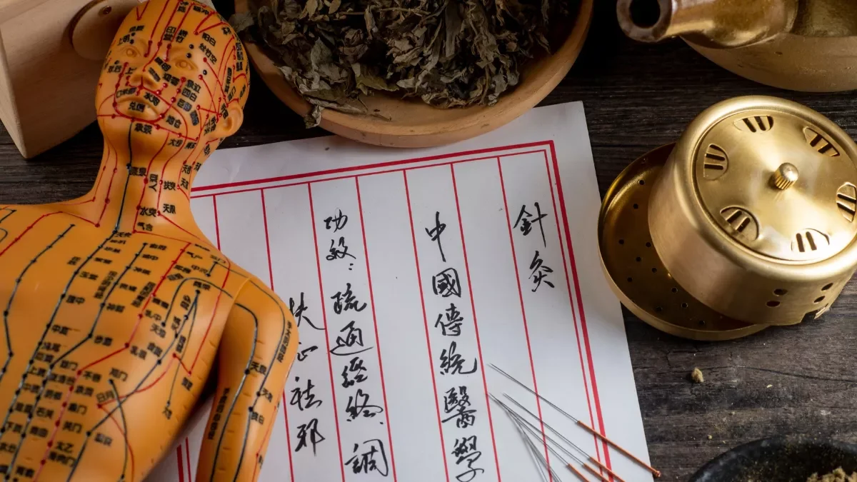 Une poupée anat ancienne avec des points d'acupuncture peints dessus, une table en bois avec du papier, une assiette contenant des herbes séchées, une horloge en laiton et un document avec des caractères chinois.
