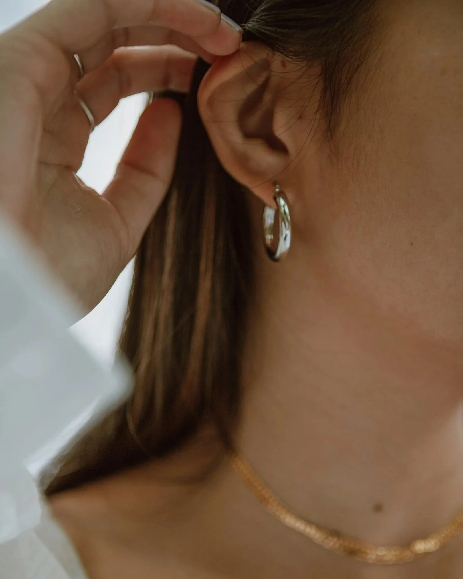 Nahaufnahme eines Ohrs mit silberner Ohrring und Halskette, Person berührt das Ohr.