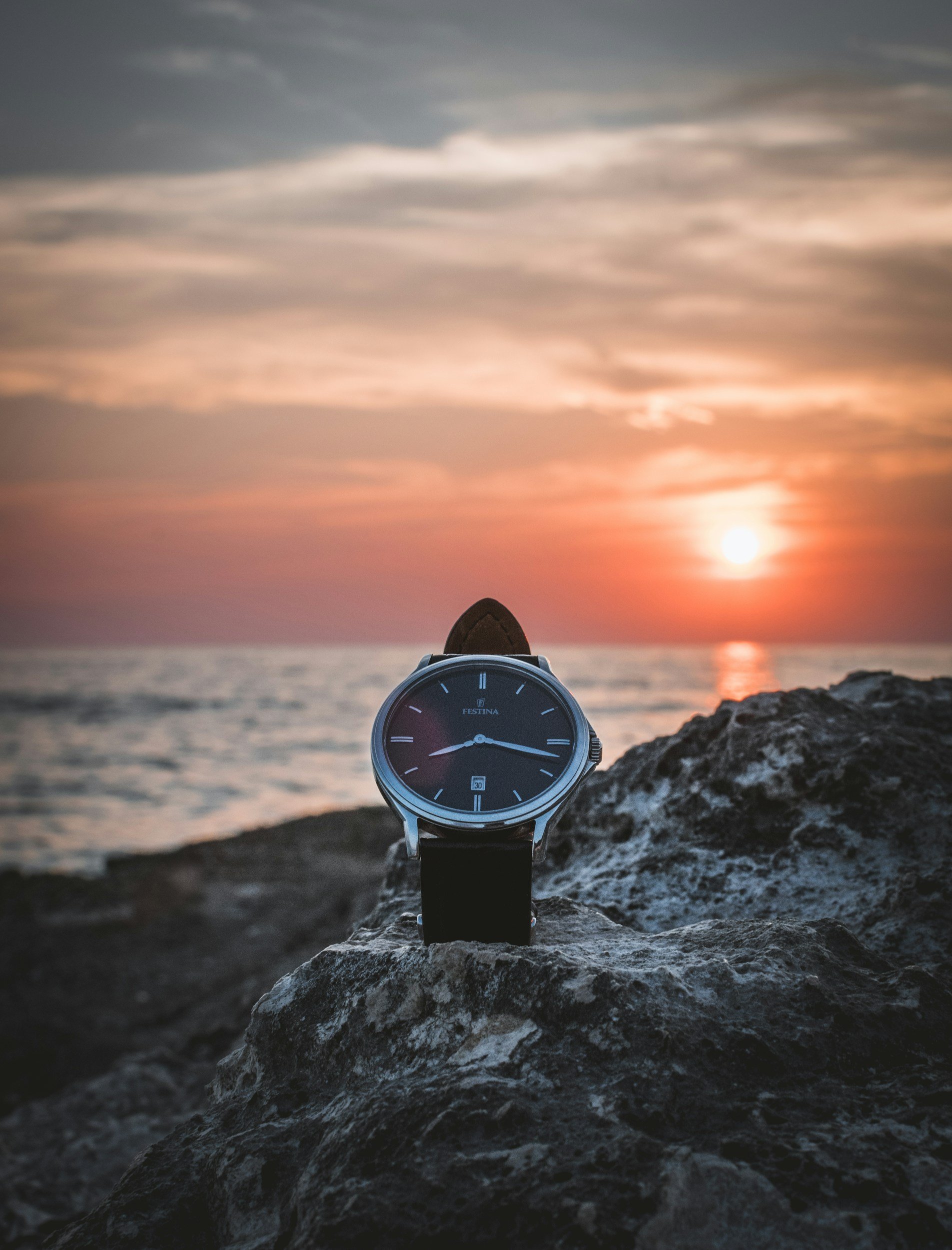 Eine Armbanduhr auf einem Felsen am Meer während eines Sonnenuntergangs, mit wolkigem Himmel im Hintergrund.