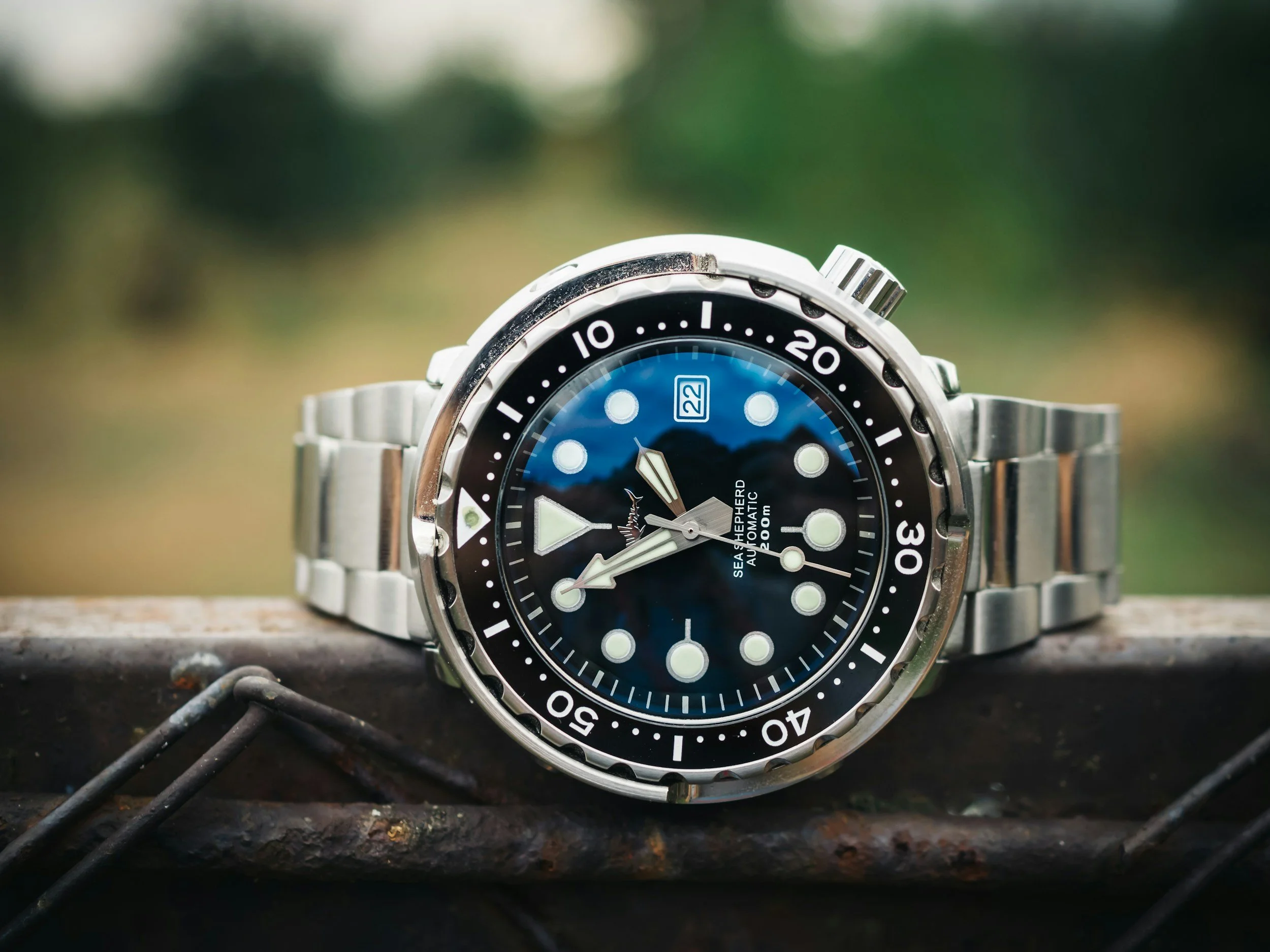 Eine Taucheruhr mit silbernem Armband, schwarzem Zifferblatt und drehbarem Keramiklünette, die auf einer rustikalen Metalloberfläche liegt, mit unscharfem grünen Hintergrund.