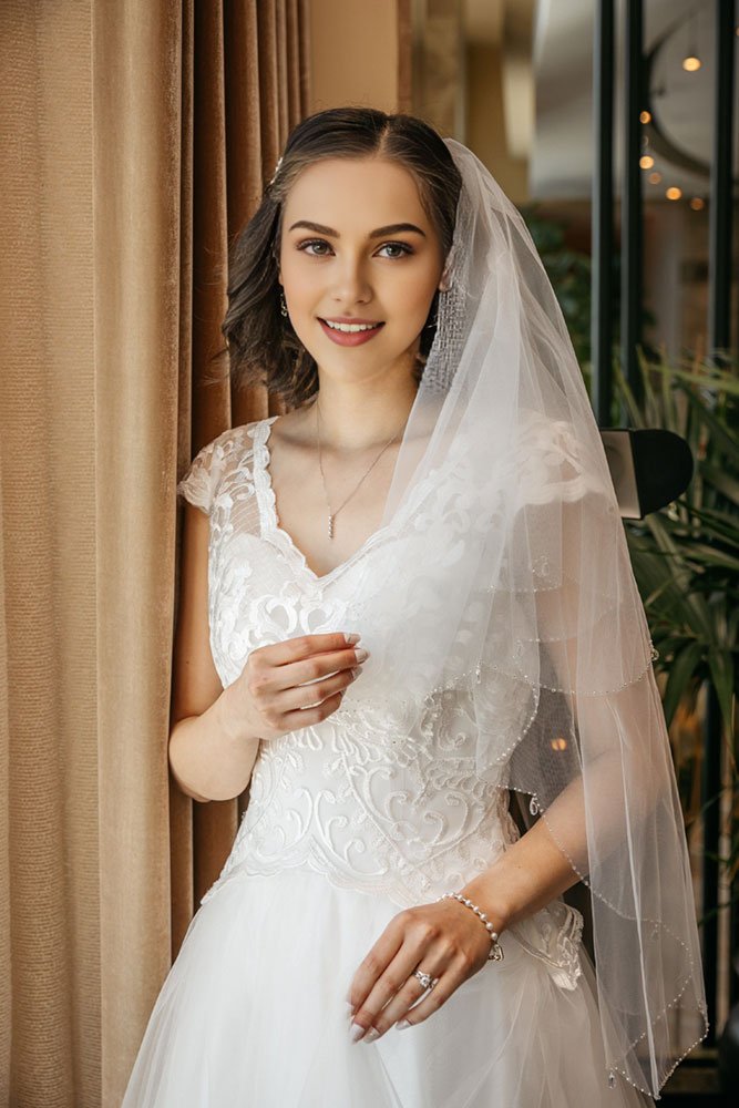 Bride wearing a short two-tier beaded bridal veil with blusher and scalloped edges, crystal-trimmed tulle.