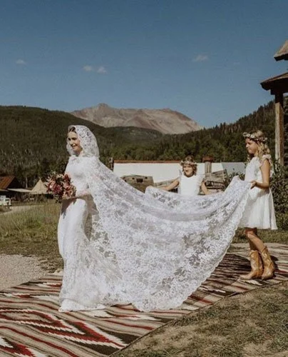 Two flower girls holding bride’s full lace hooded veil and cape, showcasing a fashion-forward 2026 veil trend.