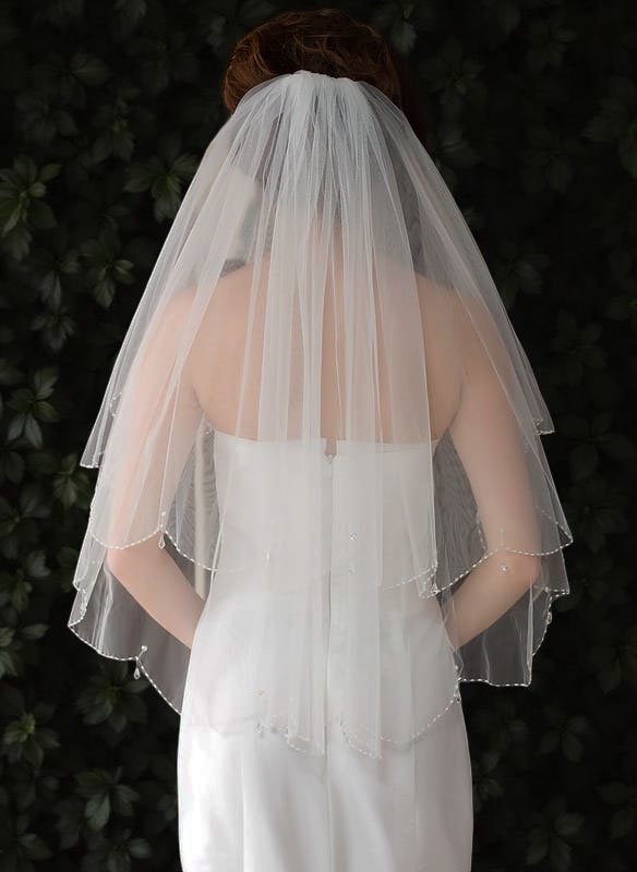 Bride wearing short two-tier bridal veil with blusher, scalloped beaded edges, and teardrop crystals adding soft sparkle.