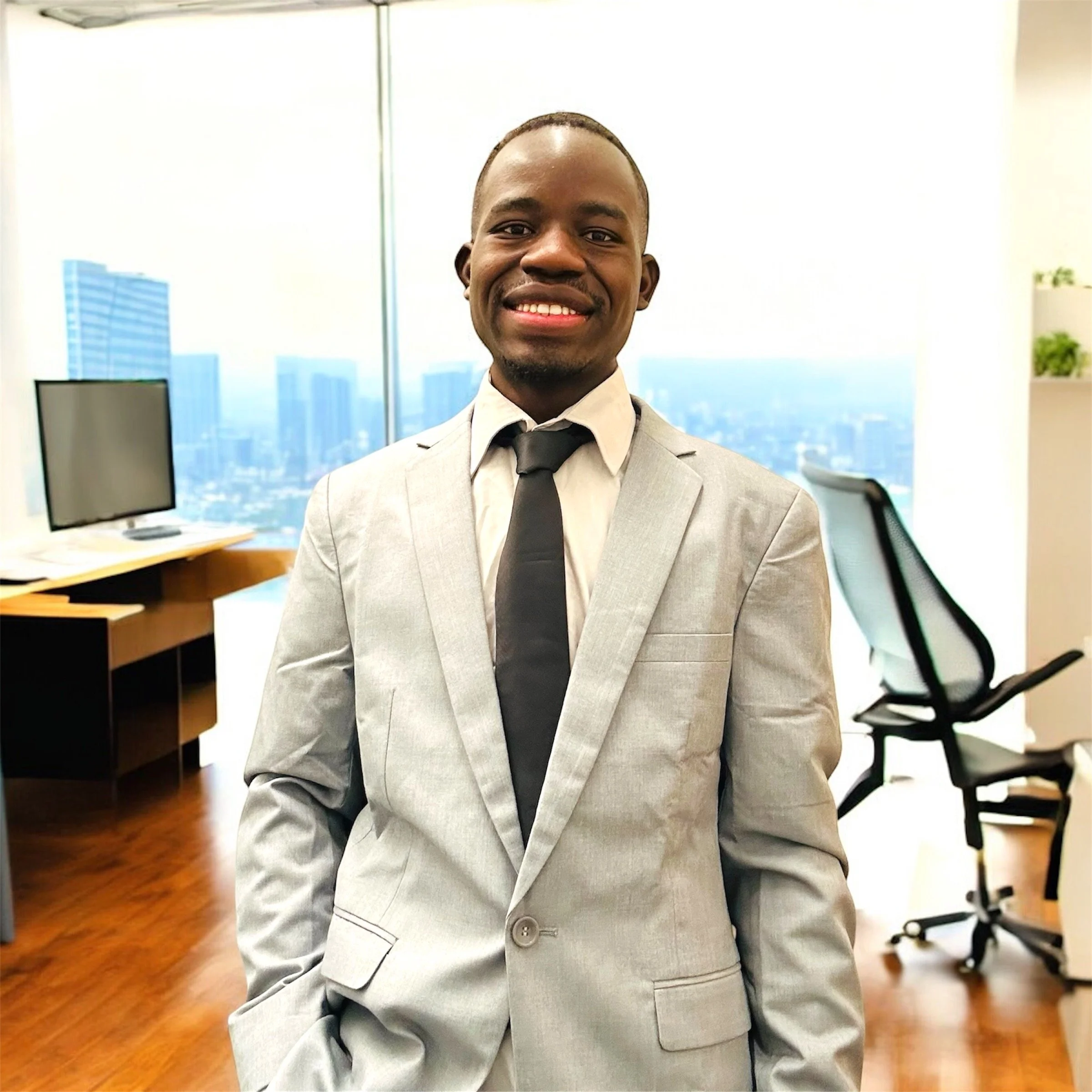 A smiling man in a light grey suit with a black tie standing in an office with large windows and a city skyline in the background.