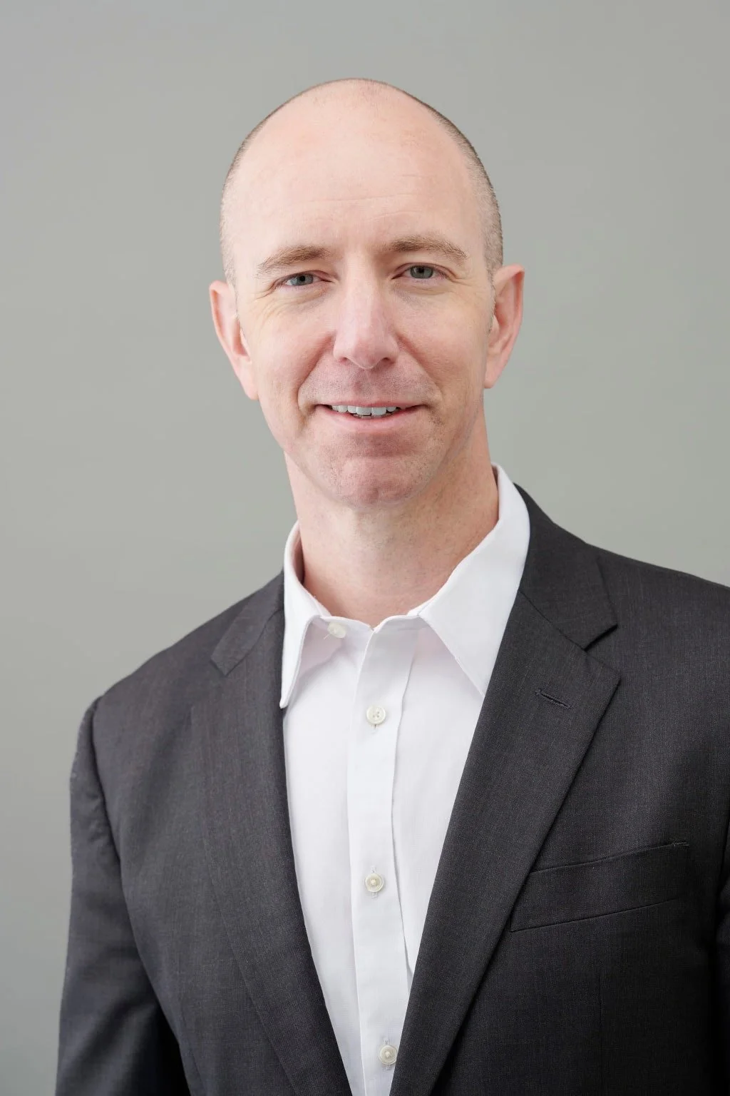 A headshot of a middle-aged man with a bald head, light skin, and blue eyes, dressed in a dark gray suit jacket and white dress shirt, smiling slightly, against a plain gray background.