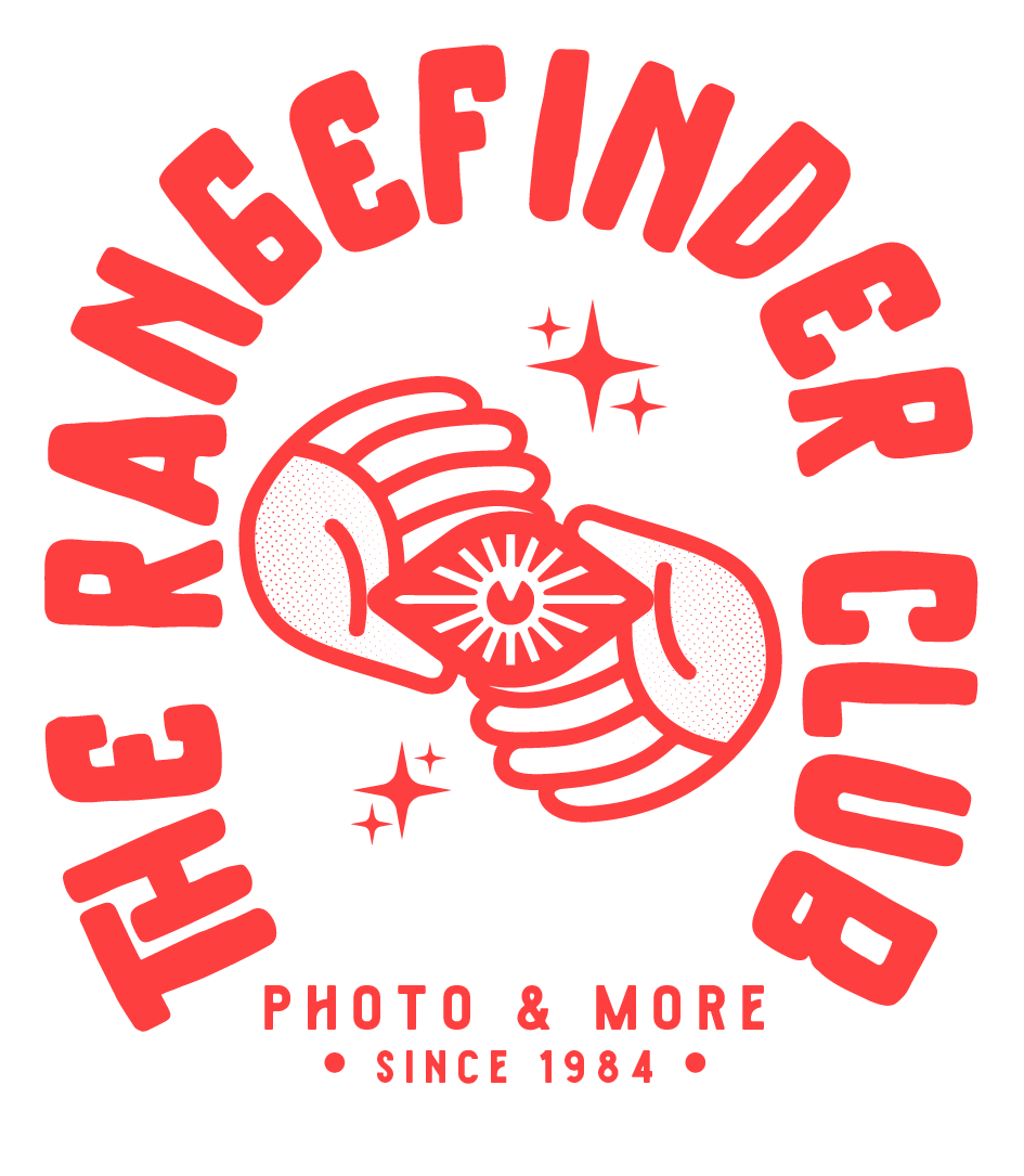 A logo with red text that reads "The Rubber Inflation Club" in a circular pattern around two intertwined hands holding a heart with a radiating line inside it. There are small star shapes around the hands. Below, in smaller red text, it says "Photo & More Since 1984."