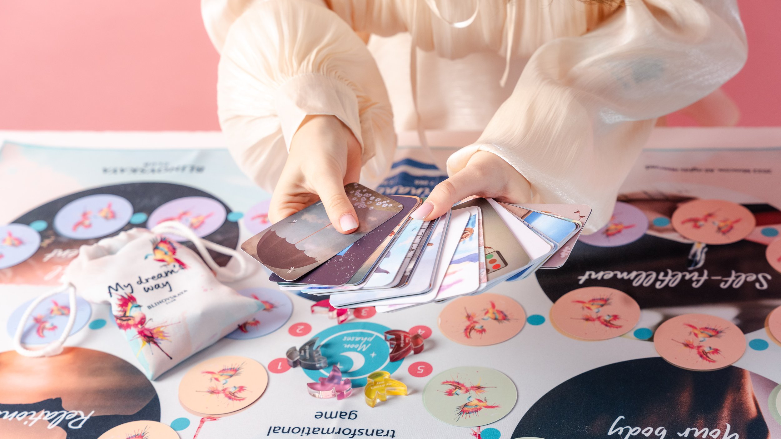 A person holds a stack of cards over a colorful board game with circular spaces and game pieces, including small figurines in different colors, on a pink background.