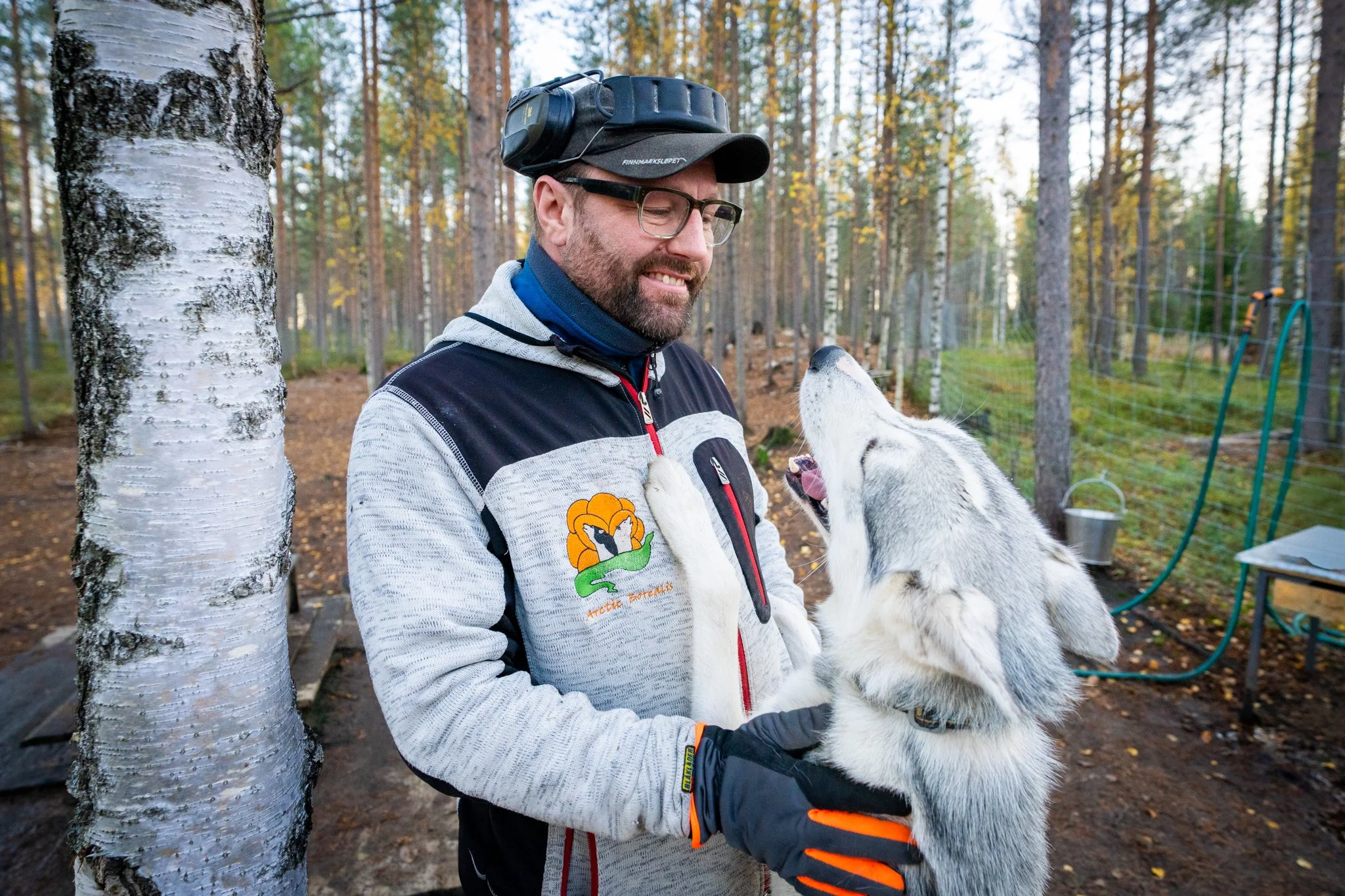 A man with glasses and beard wearing a headlamp, a hoodie, and a jacket with a lion emblem, holding a young husky puppy in a forest.