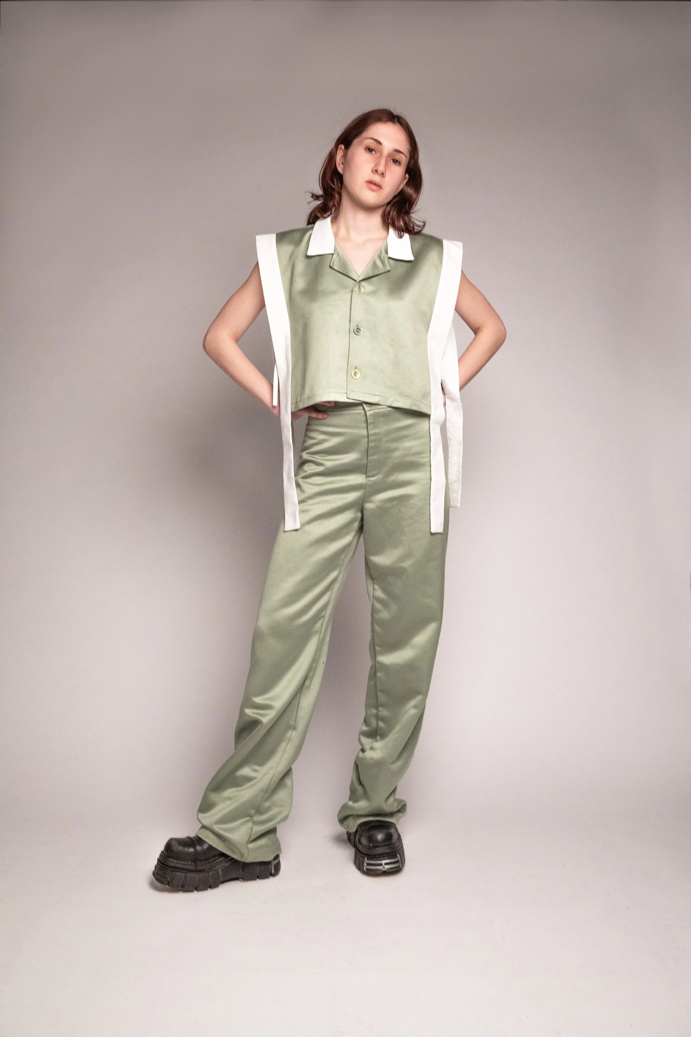 Young woman with shoulder-length brown hair posing in stylish light green satin pajama set and chunky black shoes against a plain grey background.