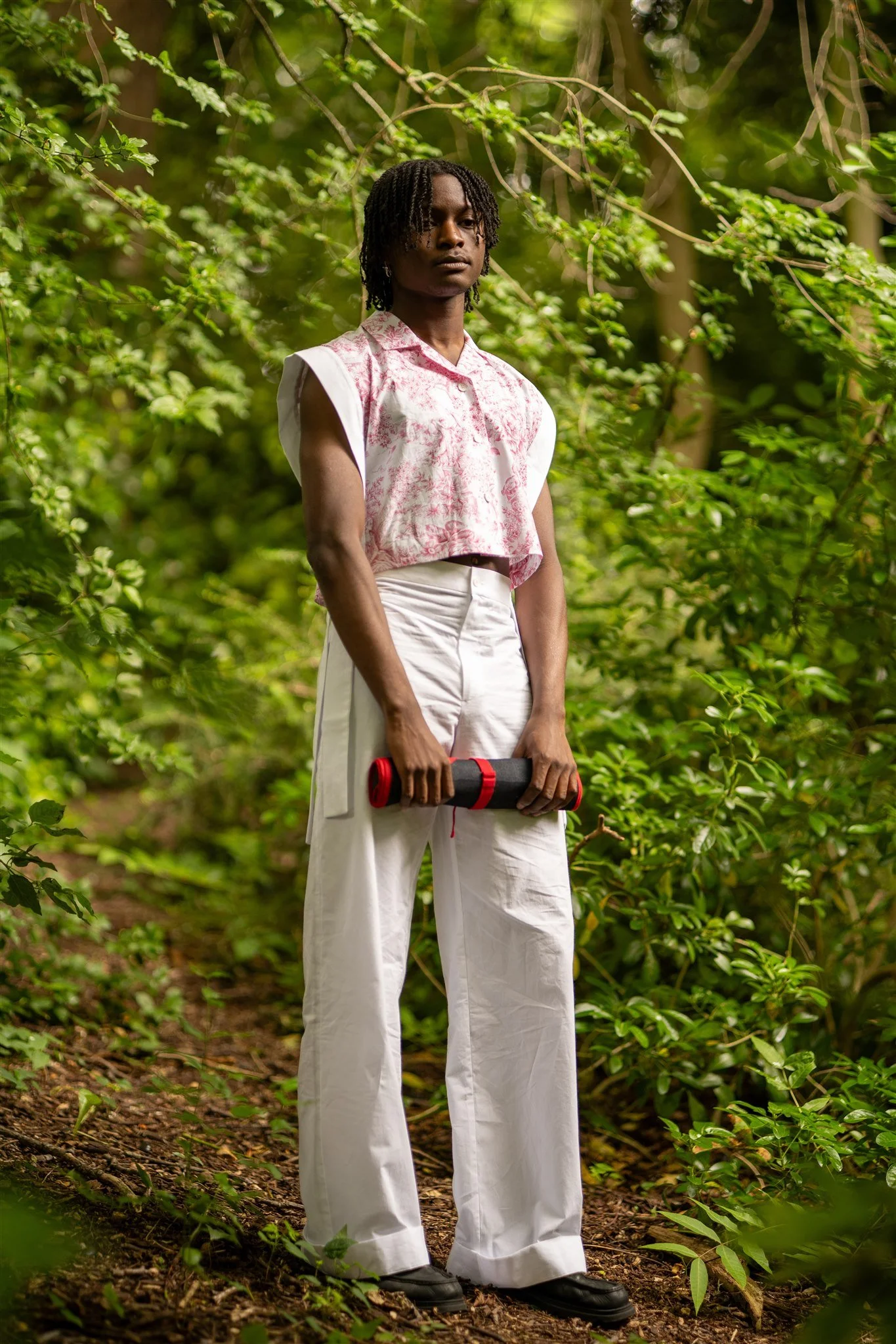 A young person with dark curly hair standing in a lush green forest, holding a rolled-up yoga mat with both hands, wearing a pink and white patterned sleeveless shirt, white wide-legged pants, and black shoes.